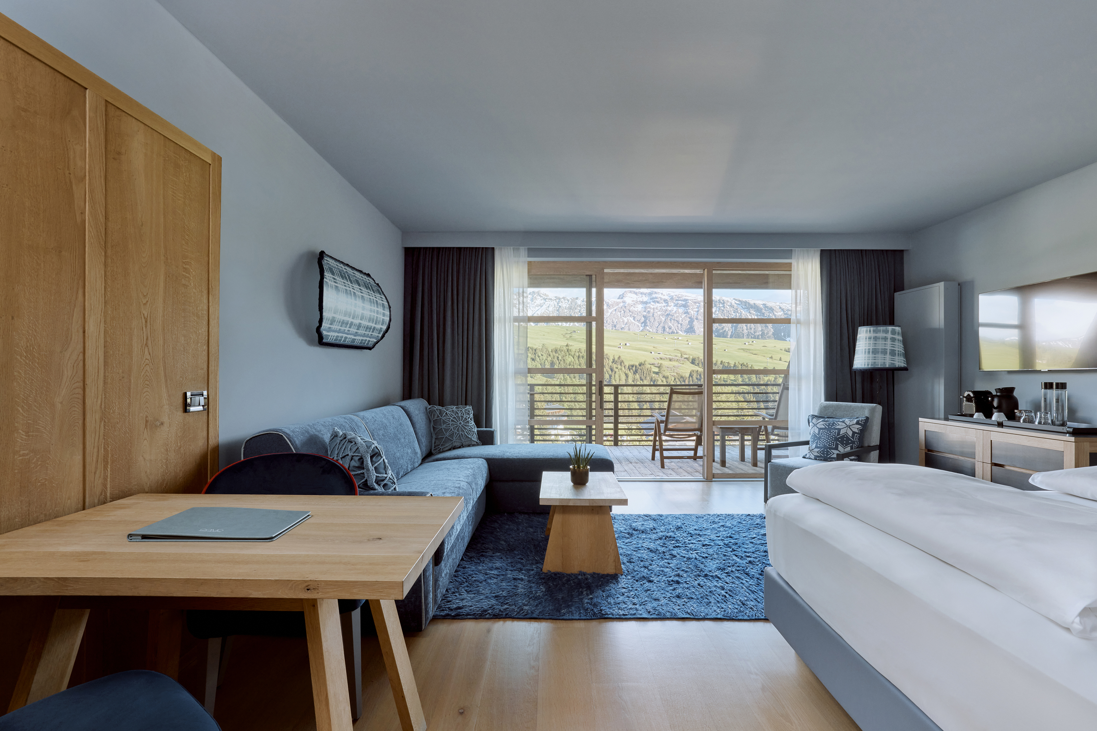 A modern hotel room with a large window offering a view of green hills, featuring a bed, sofa, wooden furniture, and blue accents.