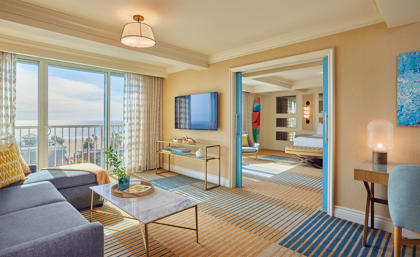 A living room of a hotel suite with a view into the bedroom and window views of a blue sky. The room is decorated in cream and blue tones and features a desk, sofa and TV area.