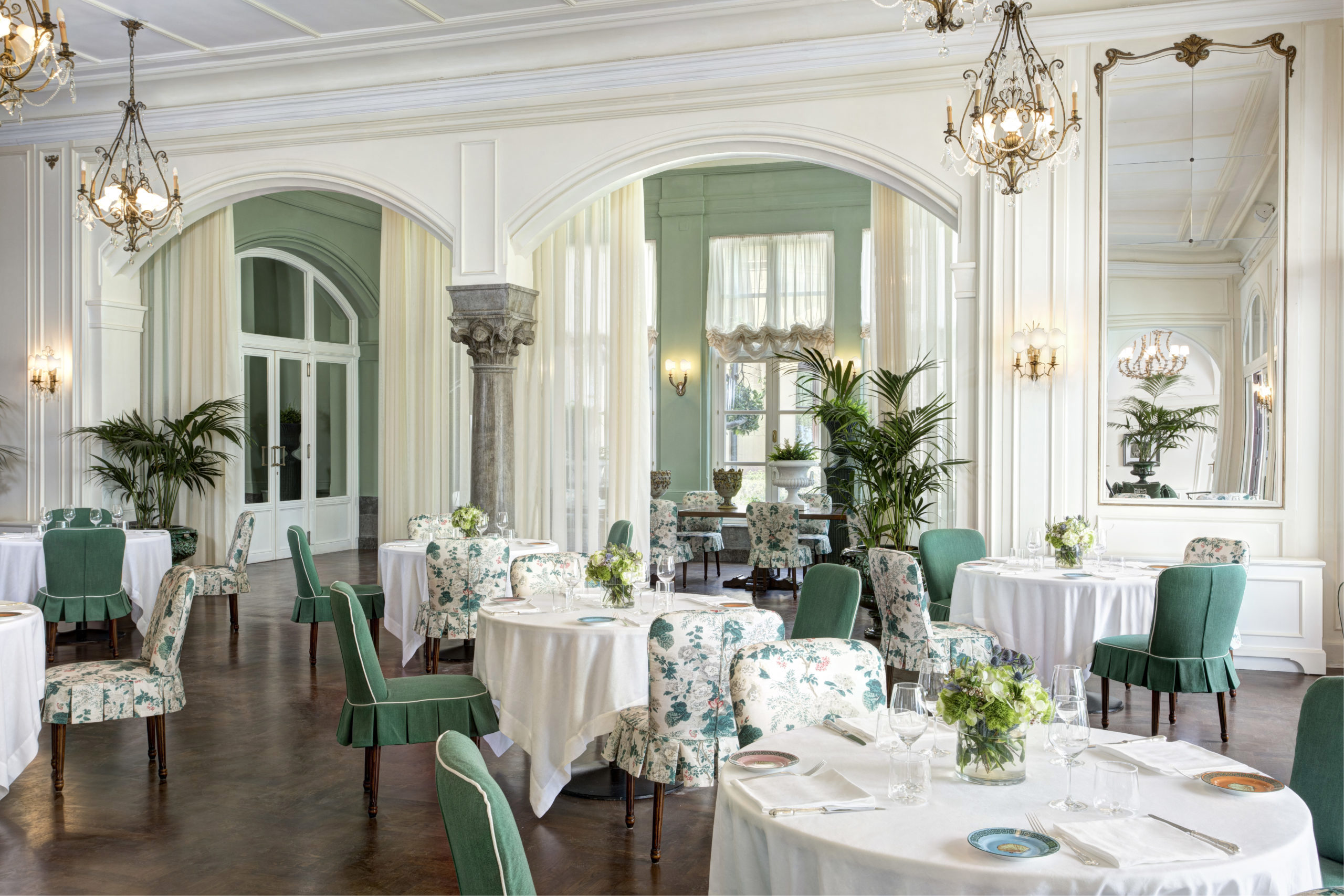 A pretty white and green dining room with multiple tables set with fine china, surrounded by green upholstered chairs, under ornate chandeliers, and large windows draped with sheer curtains.