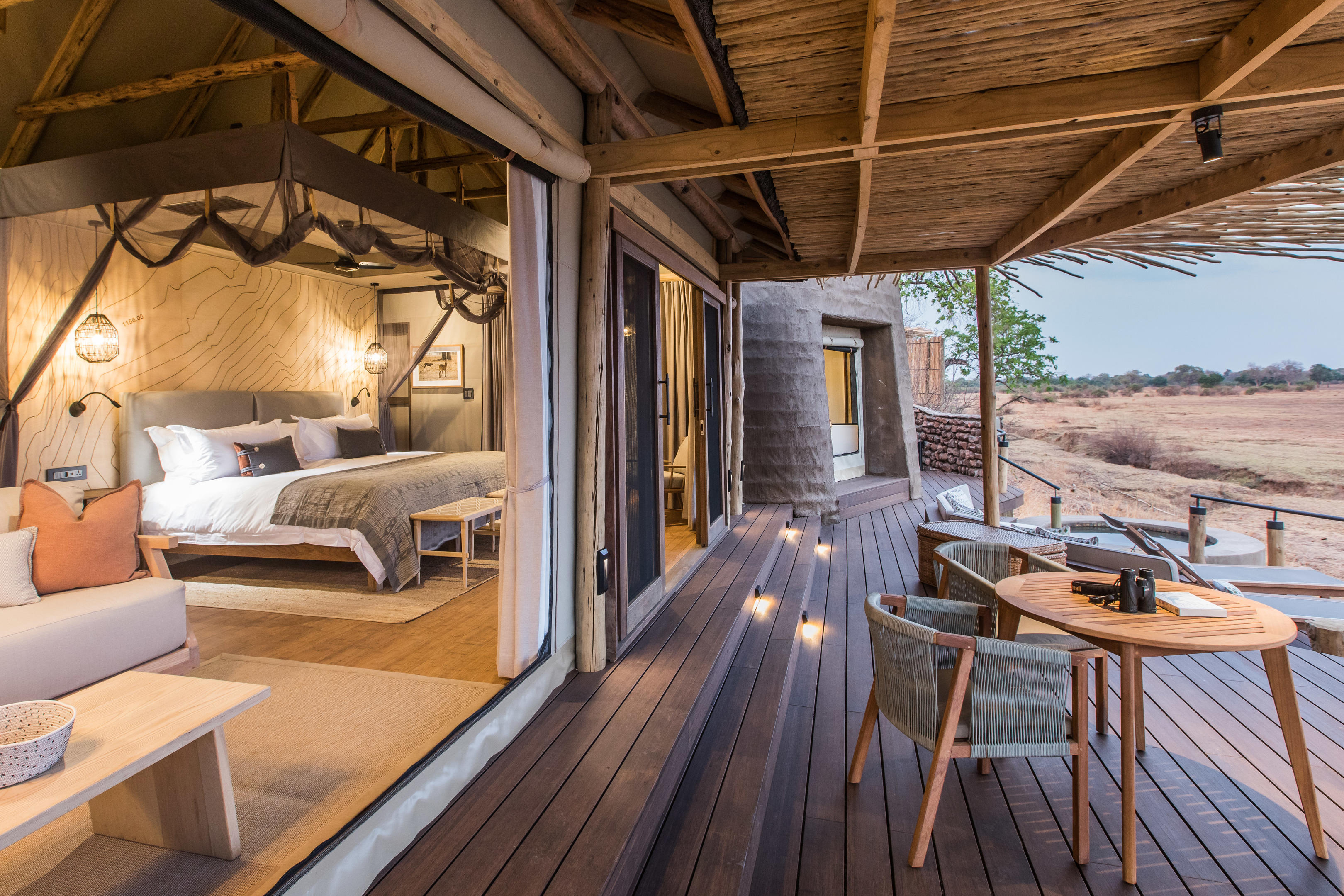 A luxurious open-air tent at Puku Ridge Camp featuring a cosy bedroom with a large bed and seating area, opening onto a spacious wooden deck with outdoor furniture and views of the surrounding wilderness