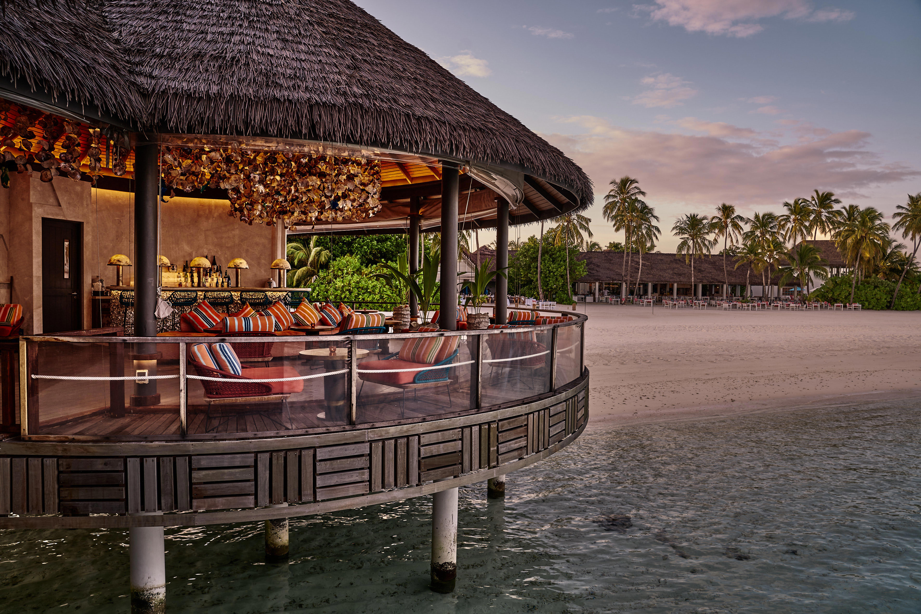 The overwater deck of the Kanusan Bar featuring colourful furniture and a statement light feature, with the beach and a thatched building behind