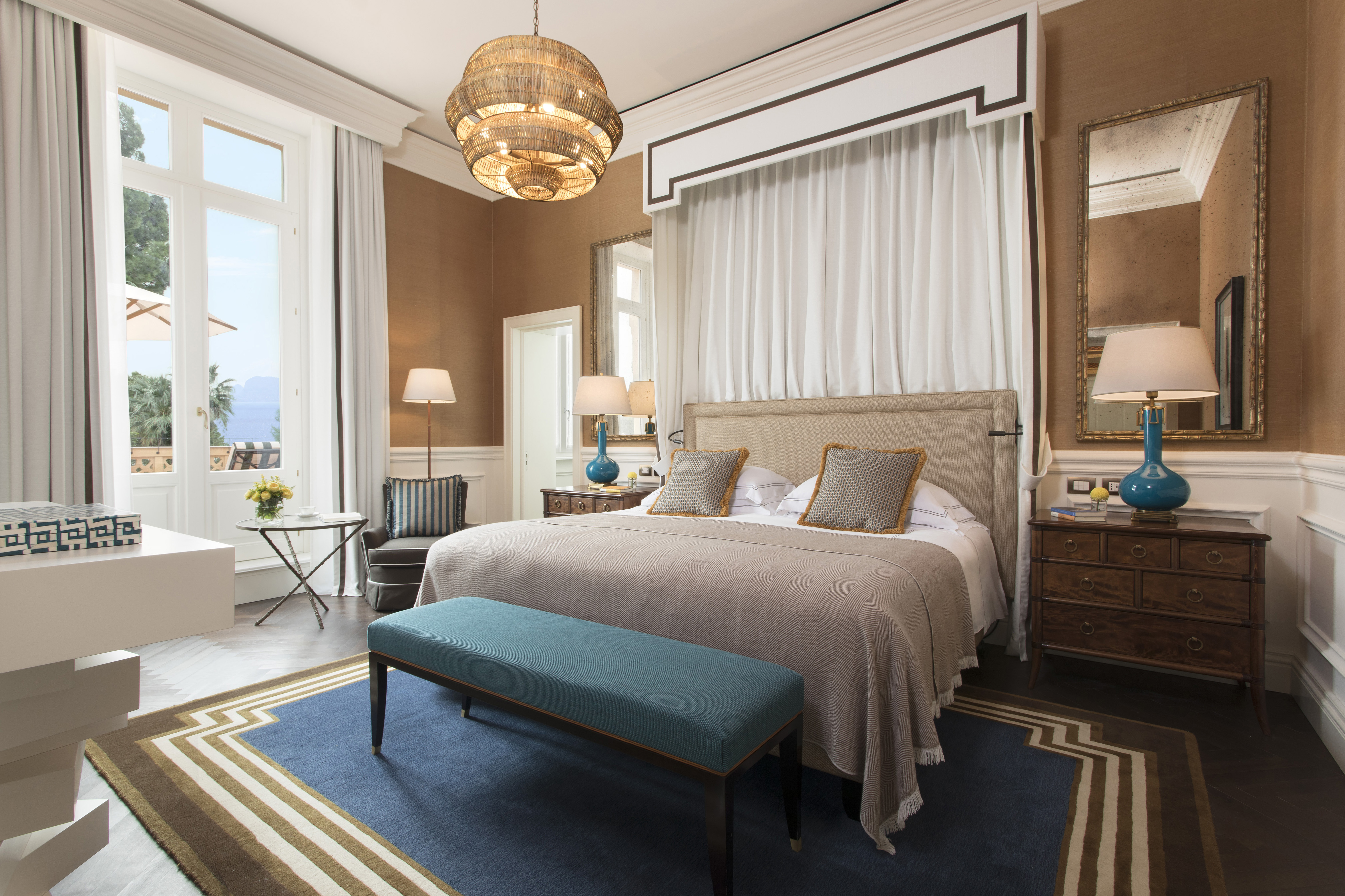 A finely decorated bedroom featuring a large bed with beige and brown bedding, a blue bench at the foot of the bed, dark wood nightstands with lamps, a chandelier, and a view of the outdoors through open windows.