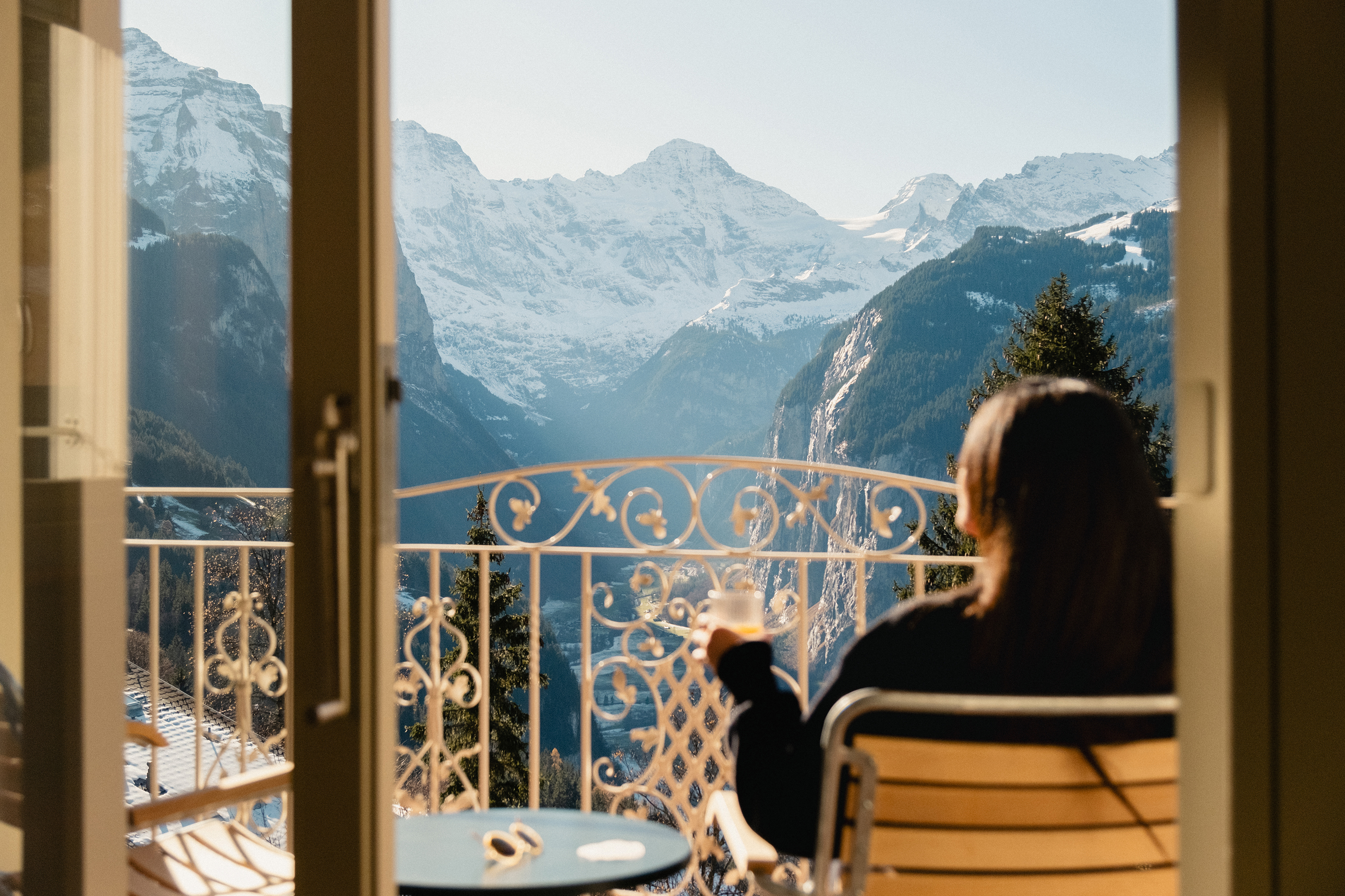 A brunette woman sitting on a balcony through open doors drinking a beverage while looking out towards a mountain view