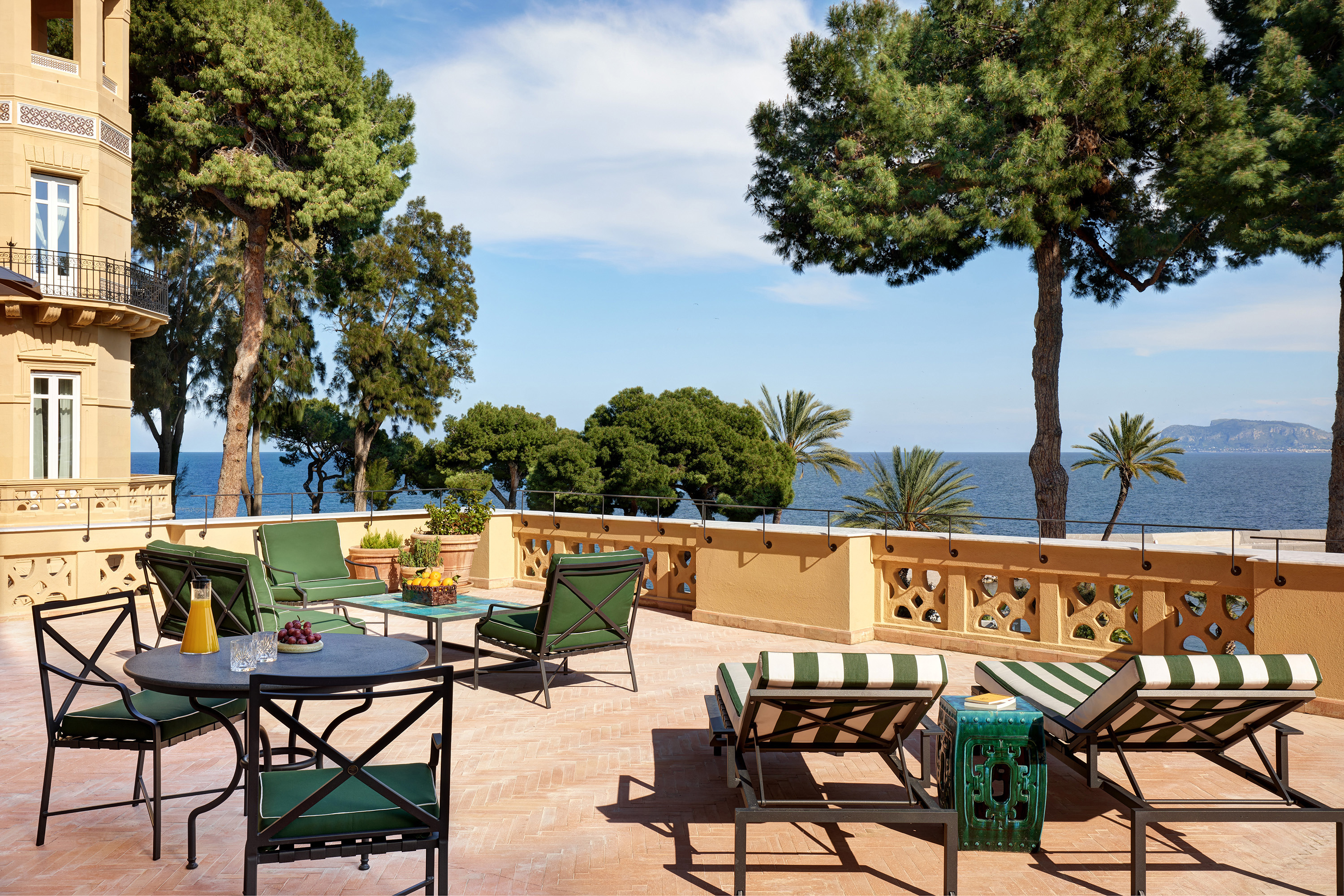A suite terrace with outdoor furniture overlooking the sea, flanked by trees and a building, under a clear blue sky.
