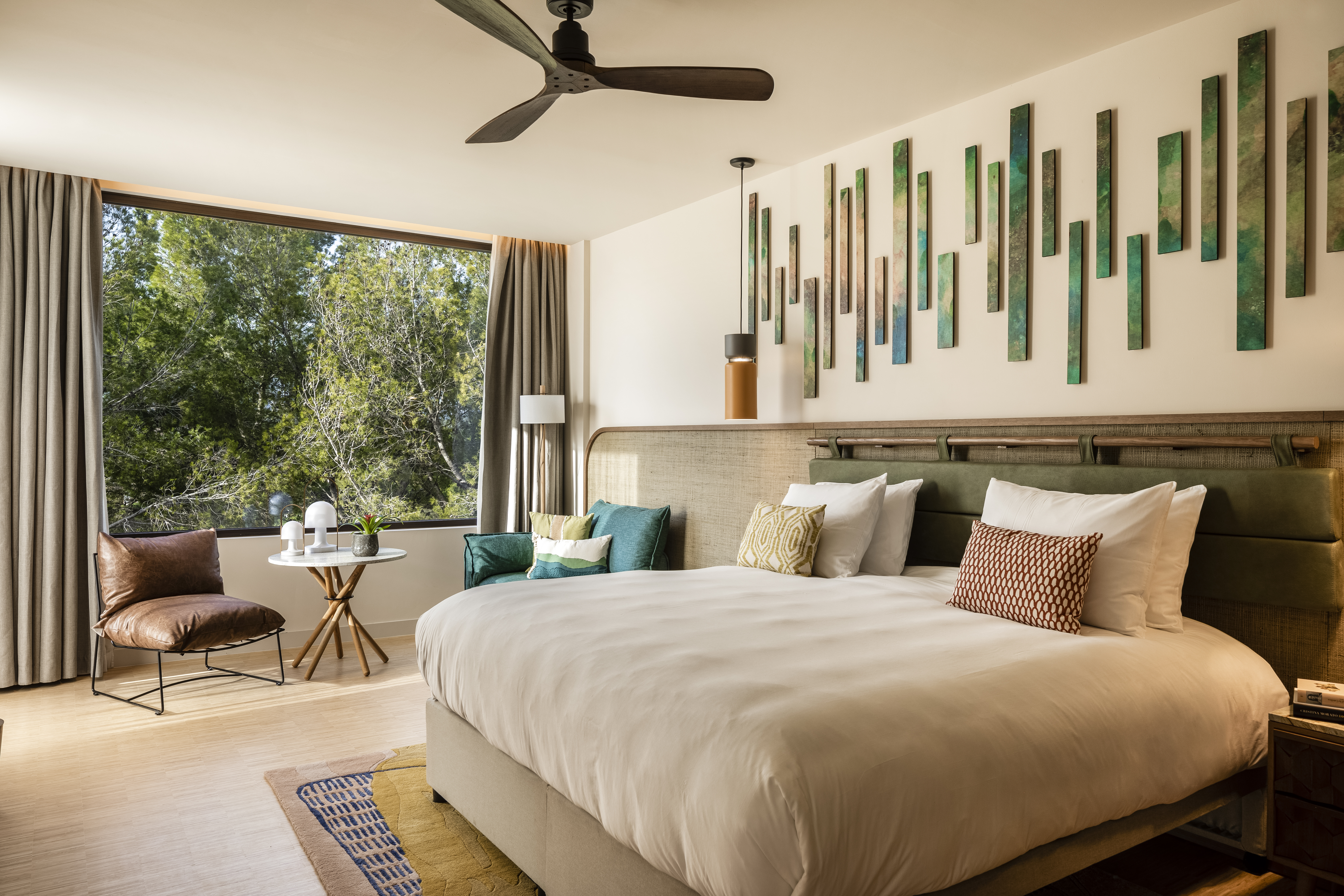 A modern bedroom with a large bed, green headboard, and multiple green wall art pieces above the bed, with a view of trees through the window