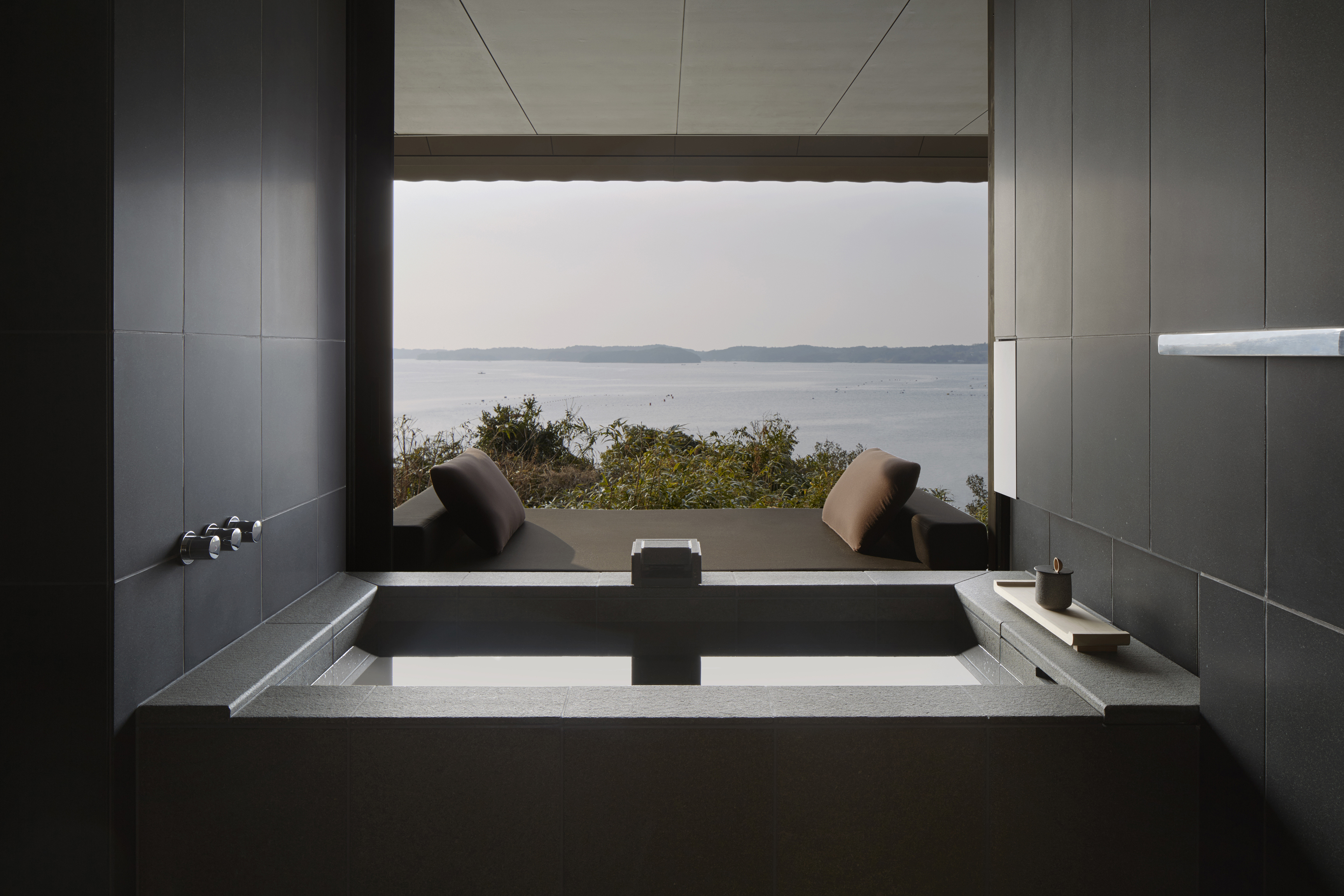 A sleek square bathtub made of dark stone with a large window looking out to a view of the ocean