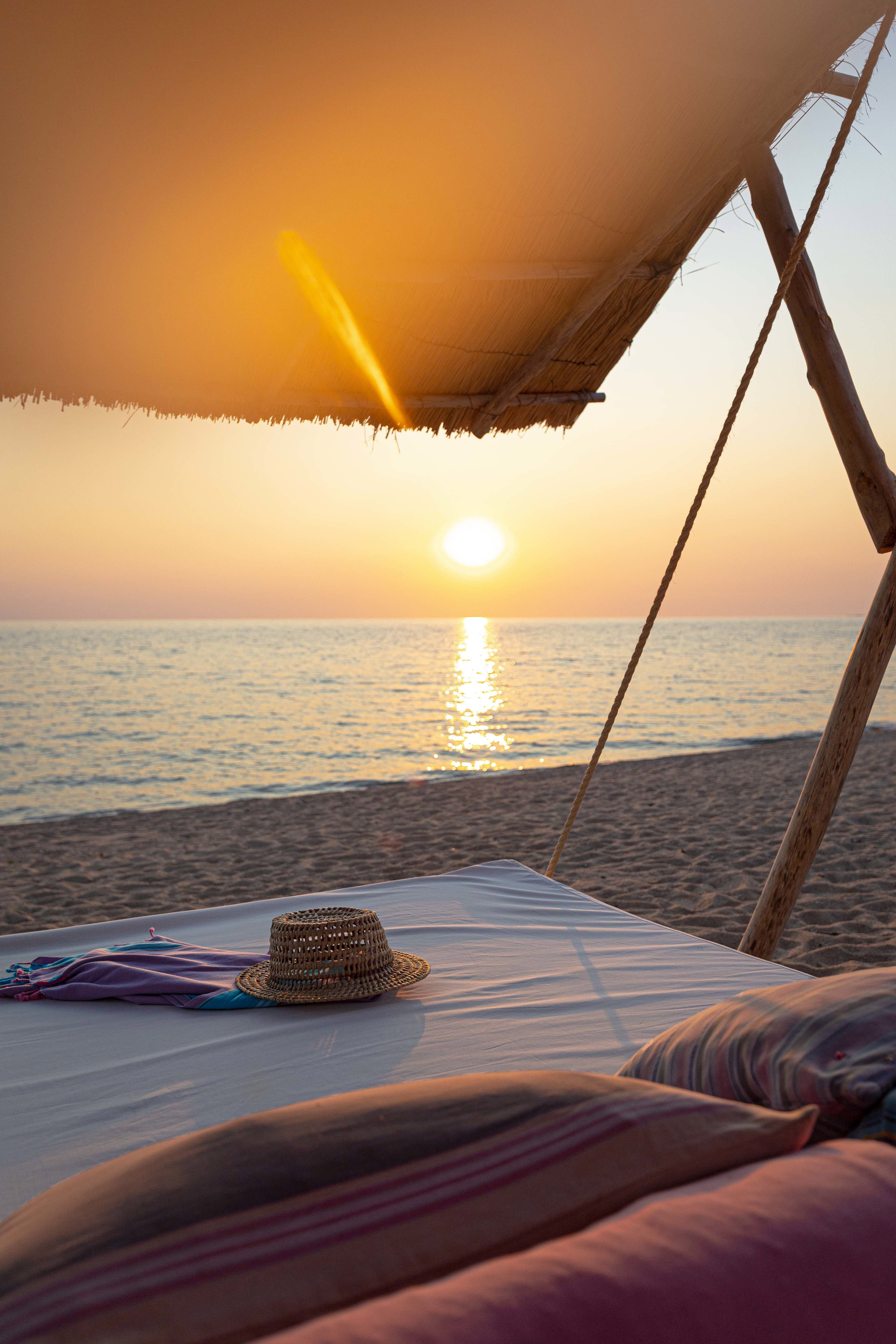 Serene beach sunset view from under a canopy with a straw hat and colorful fabric on a bed.