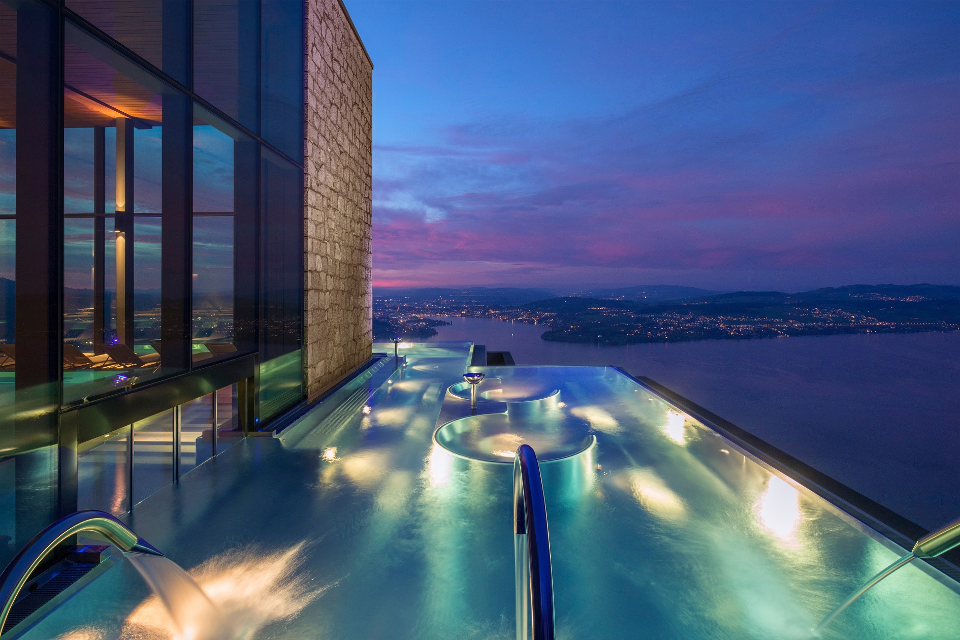 Burgenstock Hotel infinity pool with illuminated turquoise waters at twilight, overlooking a panoramic view of Lake Lucerne with mountains in the distance.