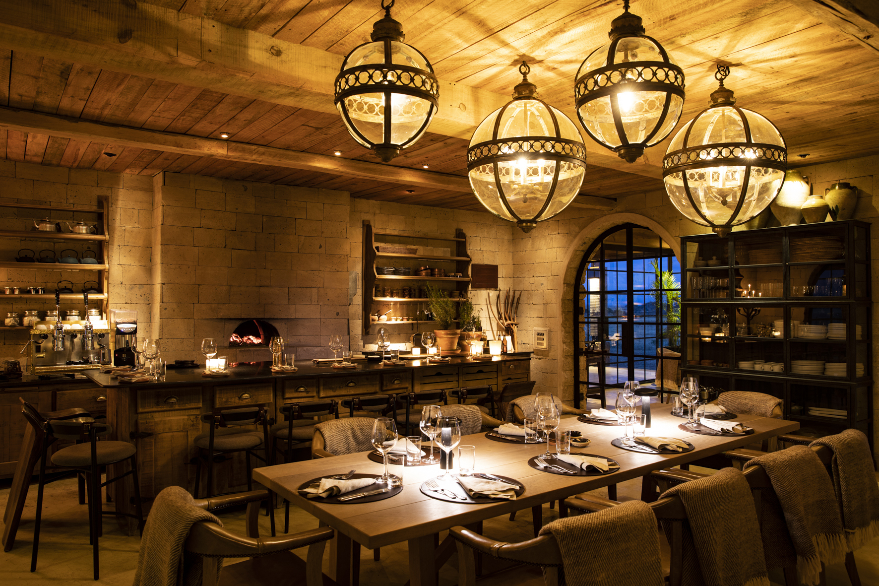A cosy dining room featuring an open kitchen and a large table set for dinner below large statement lights