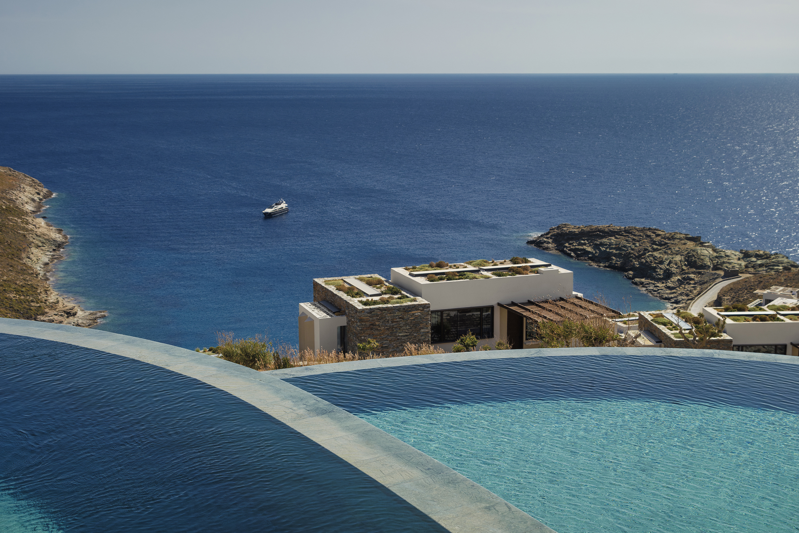 View over two infinity pools down to a white and stone building and boat floating on the sea