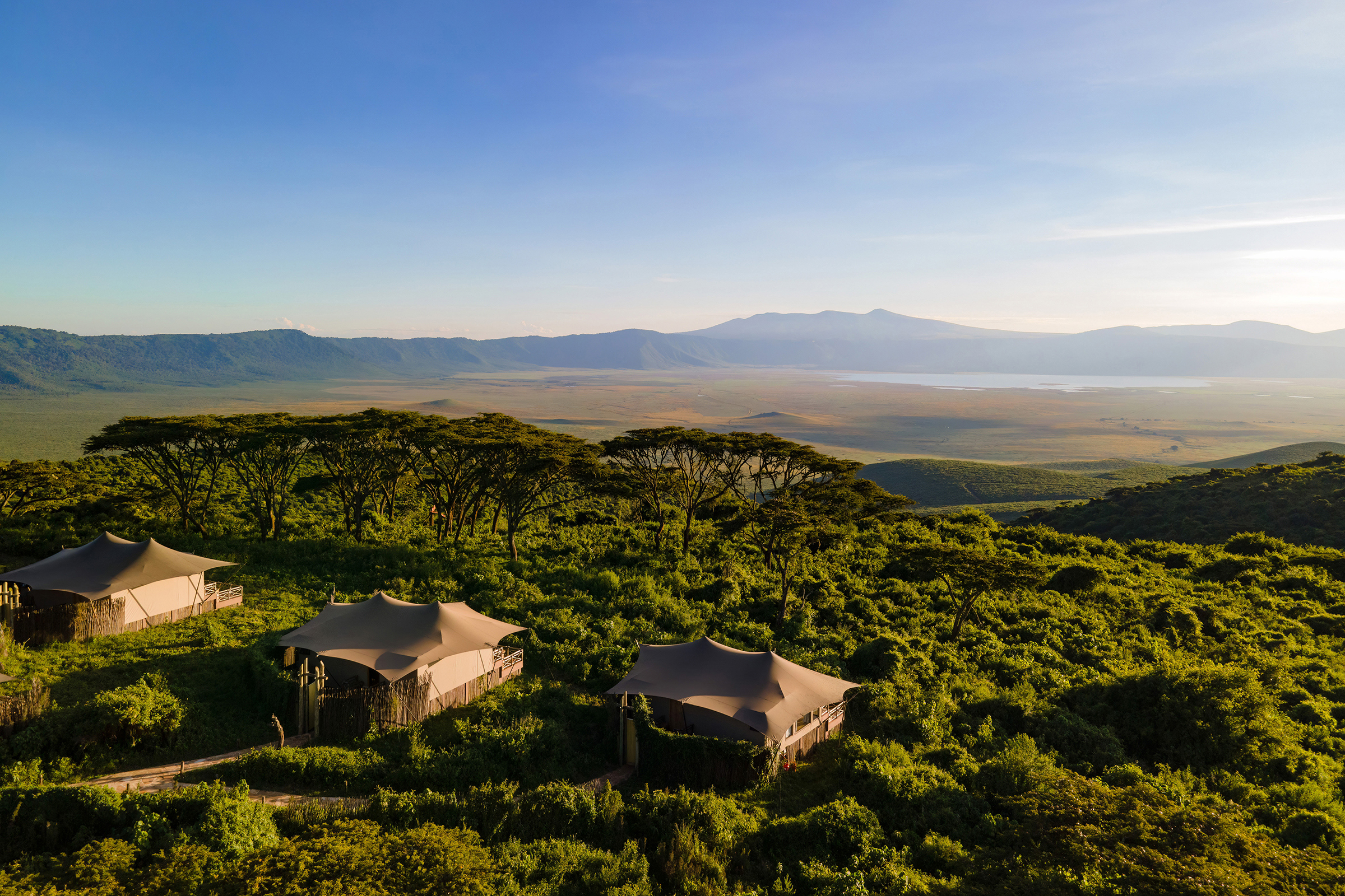 A sweeping landscape of Ngorongoro Crater with luxury tents surrounded by greenery and mountains on hte horizon under a clear sky