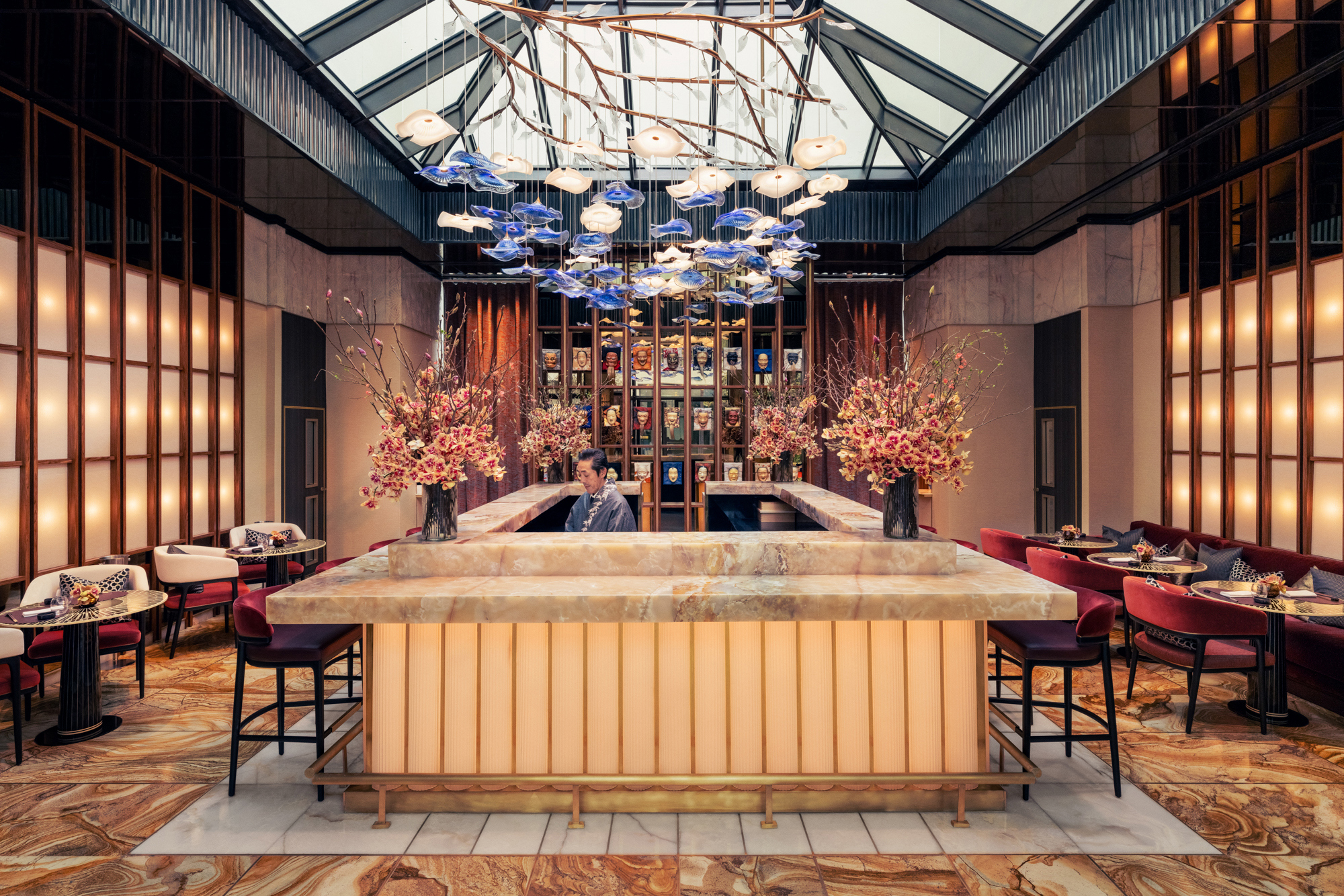 A lavish bar at Royal Mansour Casablanca featuring a person behind a marble bar top surrounded by flowers with an ornate floral light fitting above