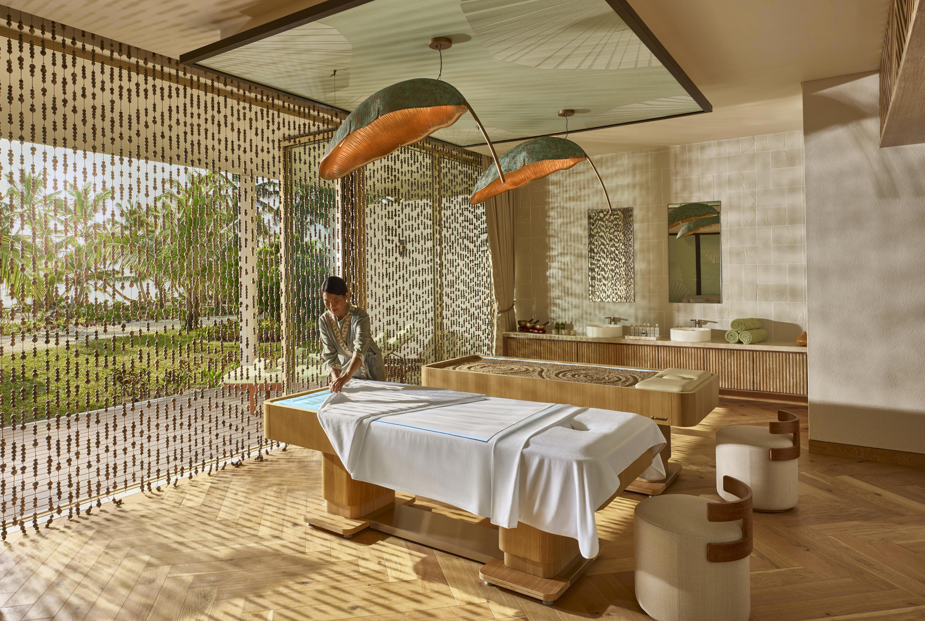  A calming spa treatment room decorated with leaf-shaped shades, a beaded curtain and natural materials, with masseuse setting up a massage bed with white towels