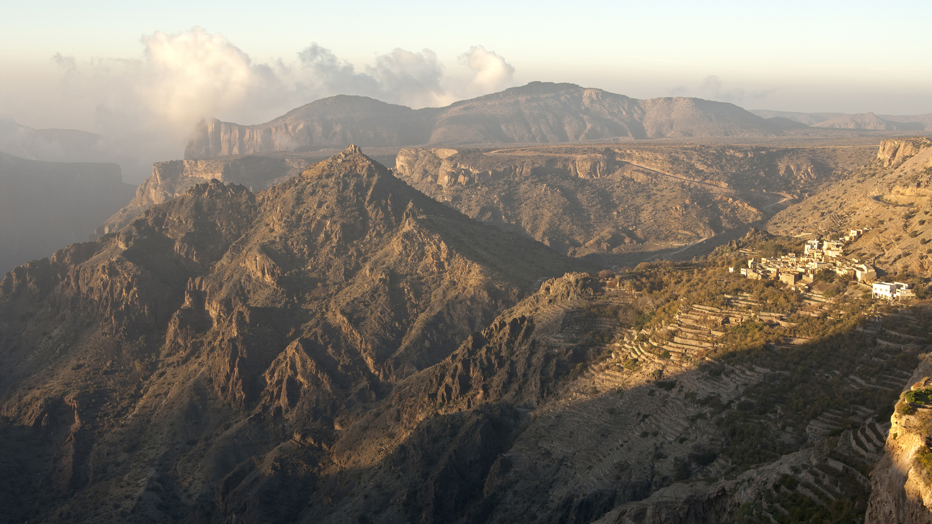 A mountainous landscape with rugged terrain and a small village on terraced slopes, shows the Saiq Plateau in Oman