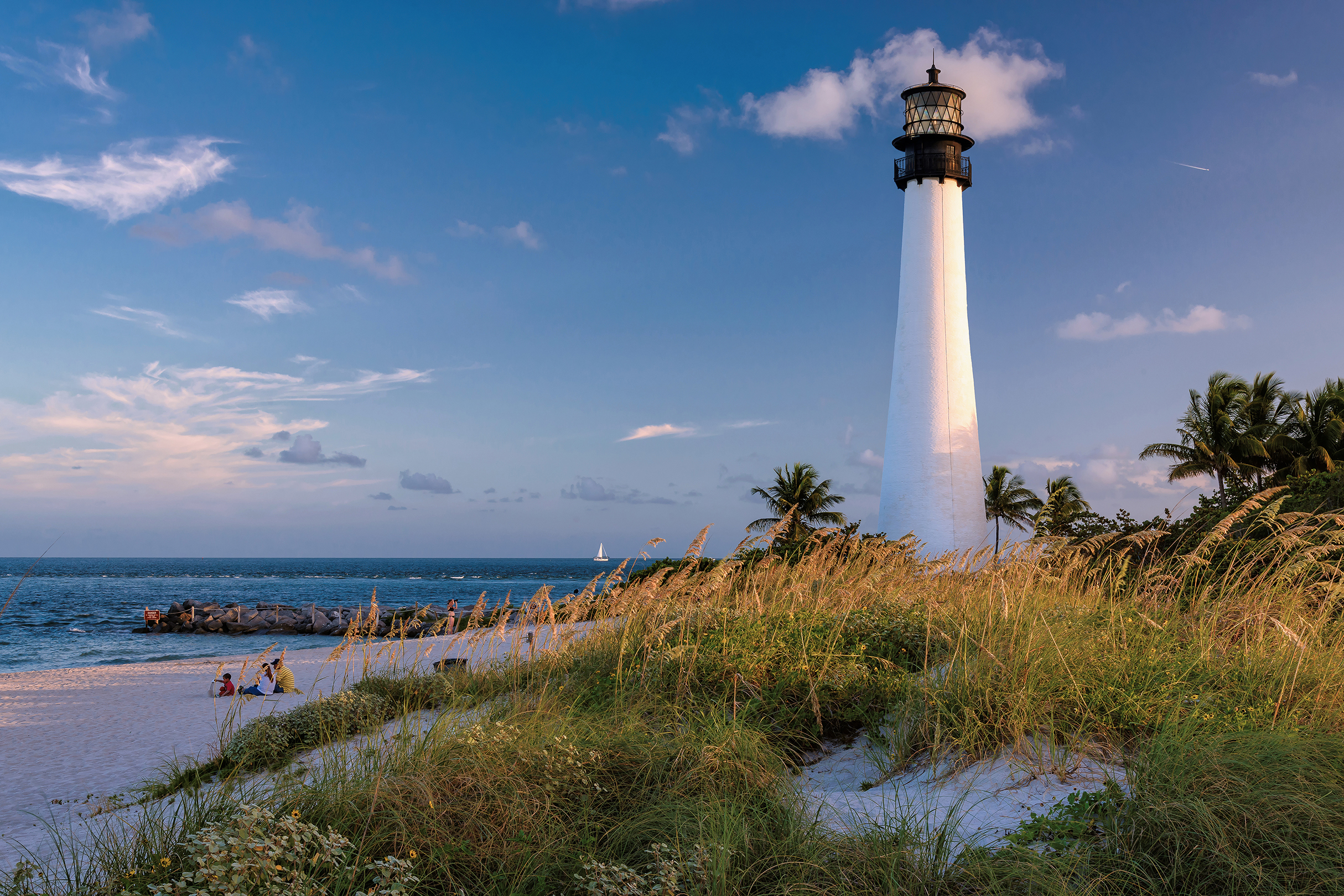 A tall, thin white lighthouse on the edge of a beach with a family sat in the sand