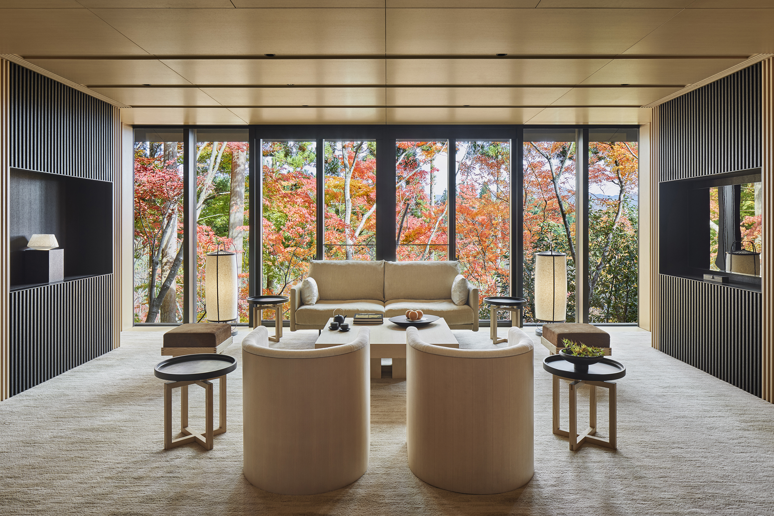 The large living room of Suite Washigamine at Aman Kyoto featuring dark wood panelled walls and pale wood furniture, with large windows that look out over trees in autumn shades