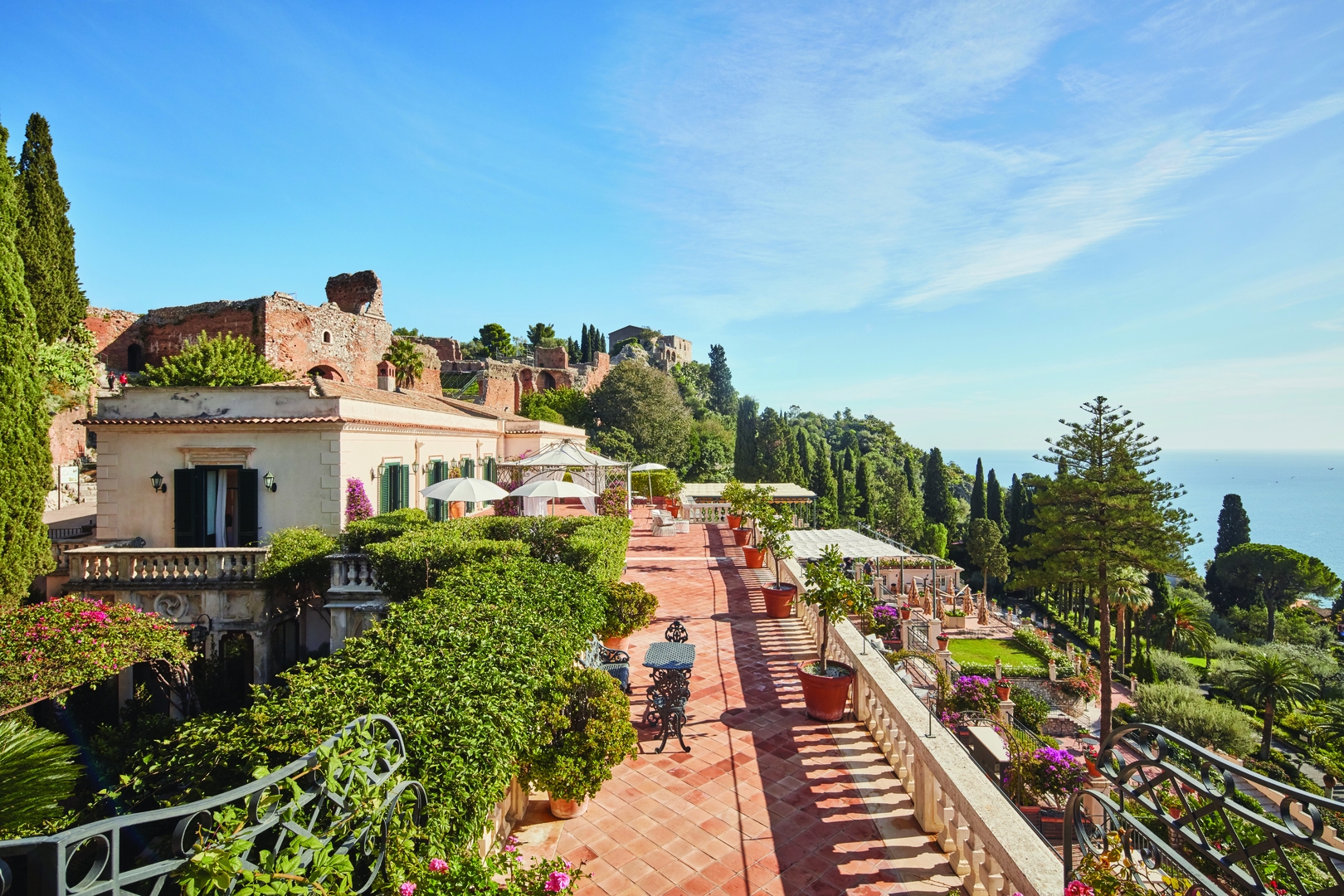 A long tiled terrace stretching between a pretty stone building and gardens leading down to the water's edge backed by a hill with a building on top at Grand Hotel Timeo, A Belmond Hotel, Taormina