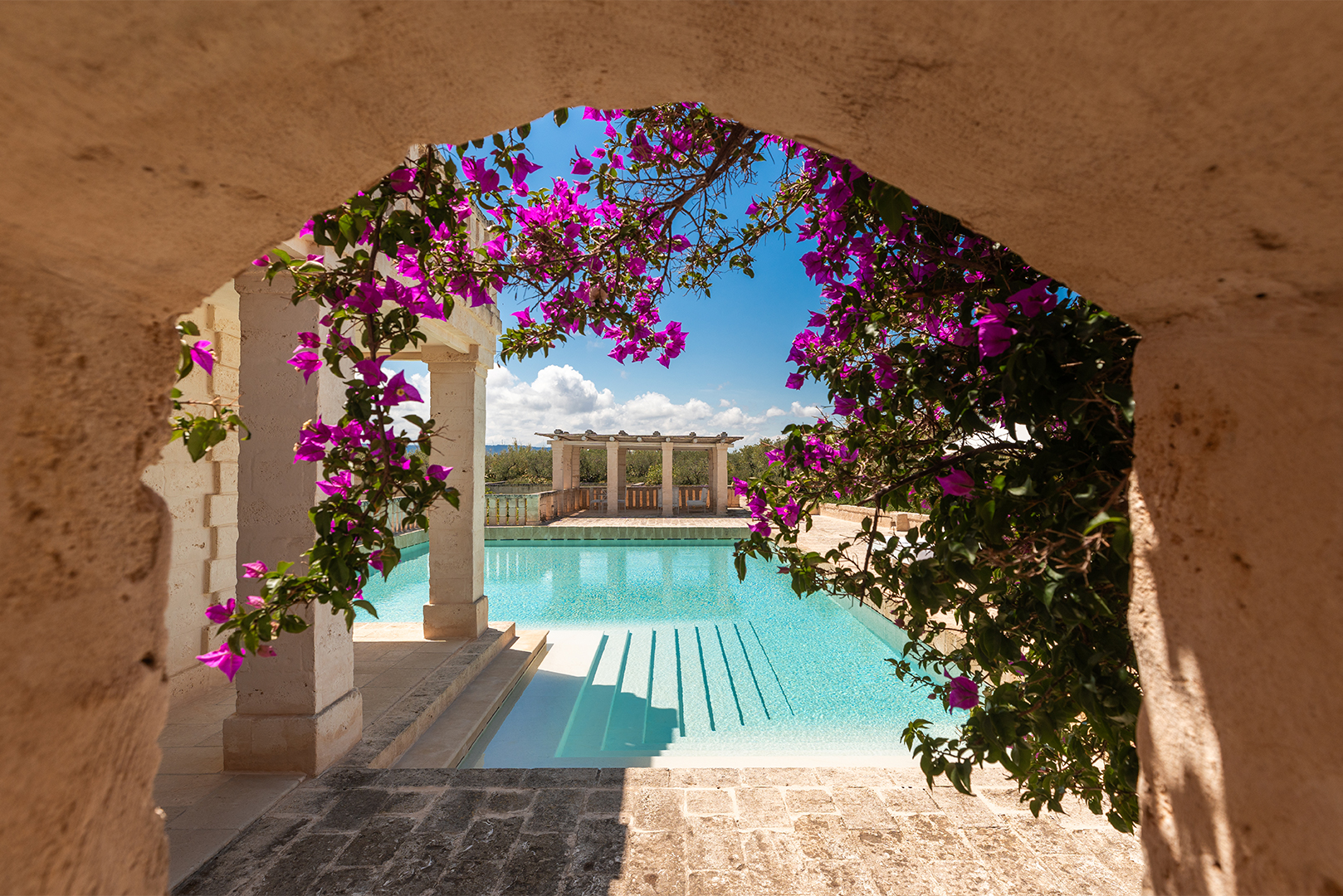 View of a tranquil pool framed by an arched stone doorway, with vibrant purple flowers hanging overhead and a clear blue sky in the background.