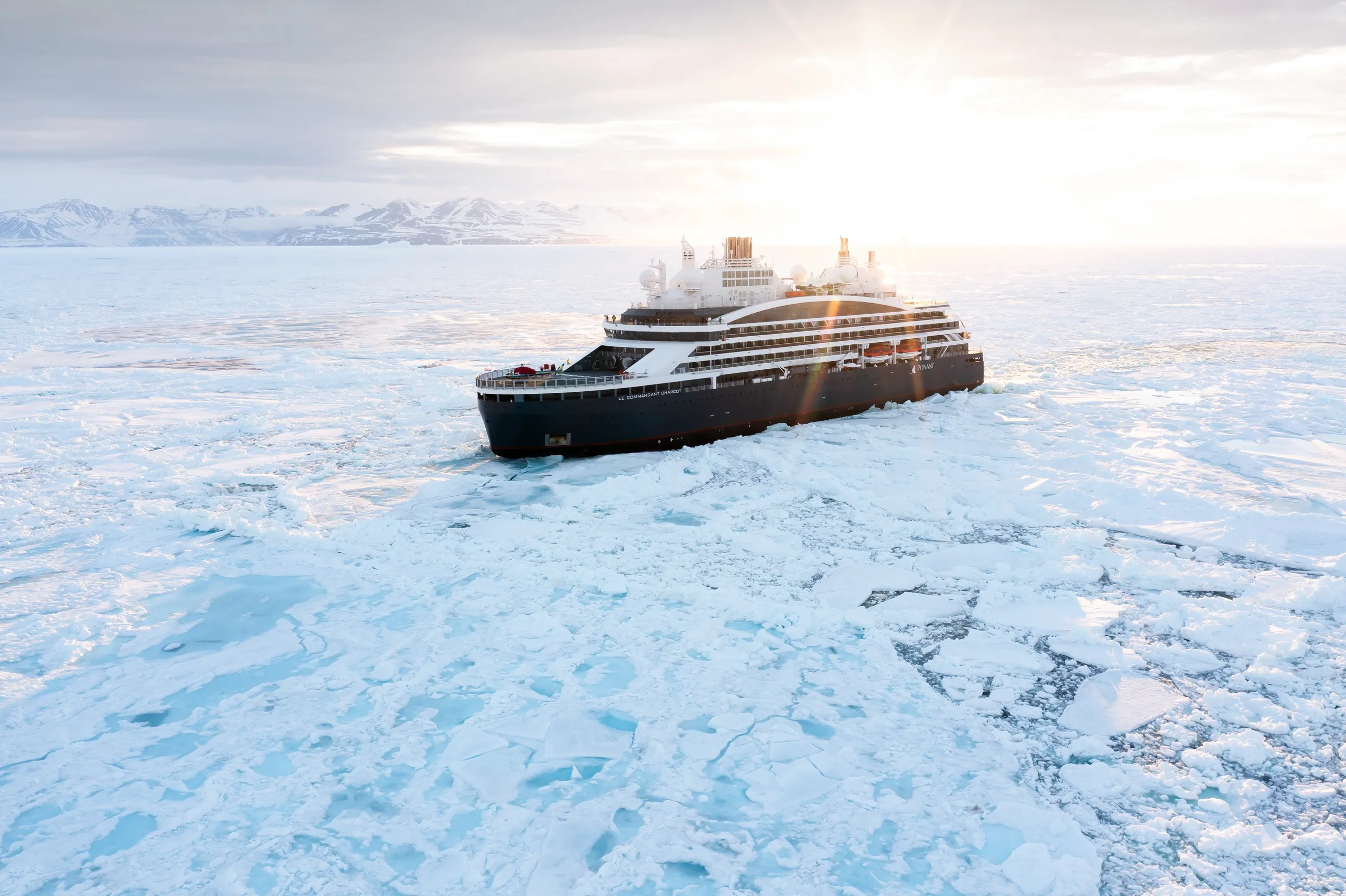Ponant expedition ship cruising through icy waters with snow-covered mountains and a low sun on the horizon.