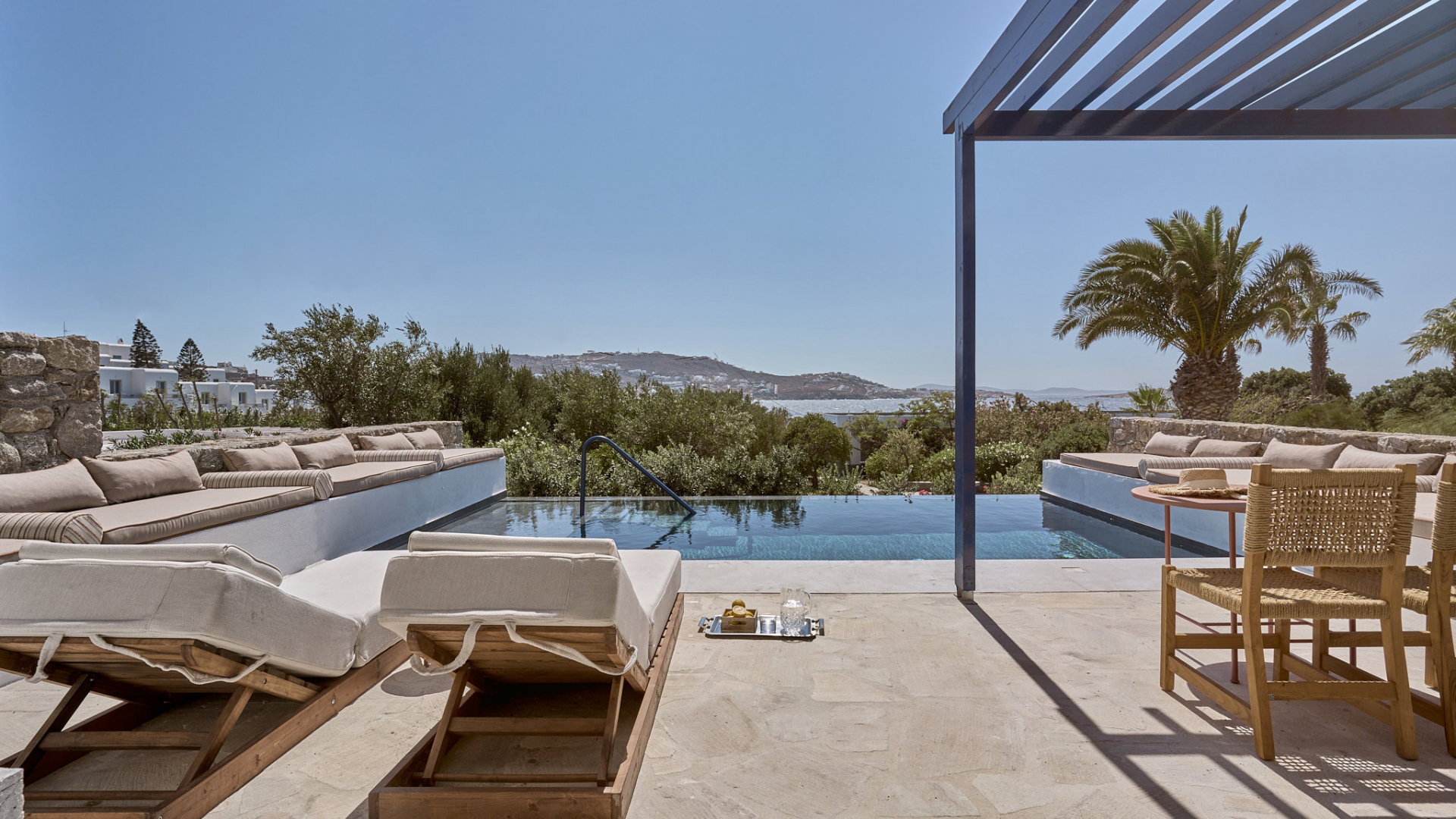 A sleek pool terrace with sun loungers and a tray of beverages beside the pool and a glimpse of a blue cabana looking out over the oceana and hills beyond