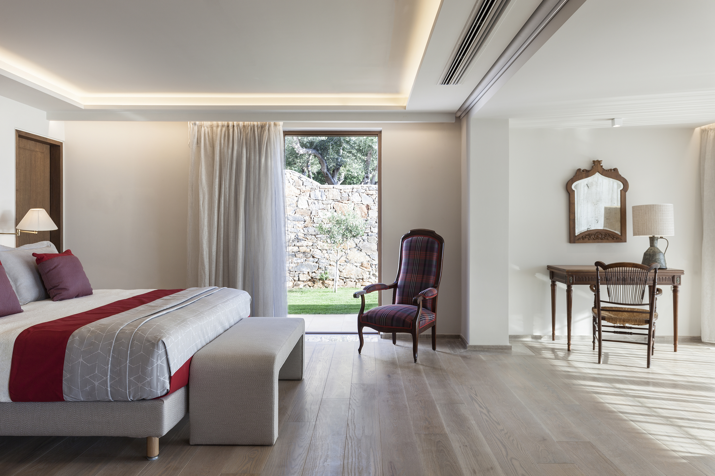 Bedroom of Minoan Palace with accent chair and bed with cream and red bedding to the left and a wooden desk and chair to the right