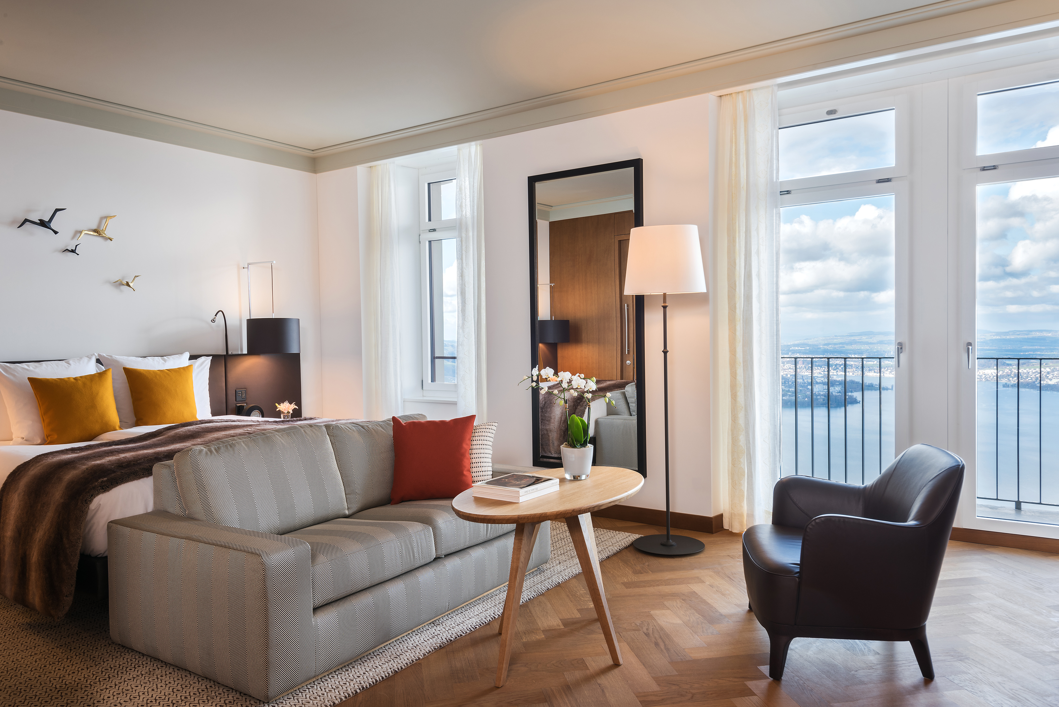 The Lake View Deluxe Room at Bürgenstock Hotel featuring a plush bed, sofa and table and chair with large windows looking out over the lake