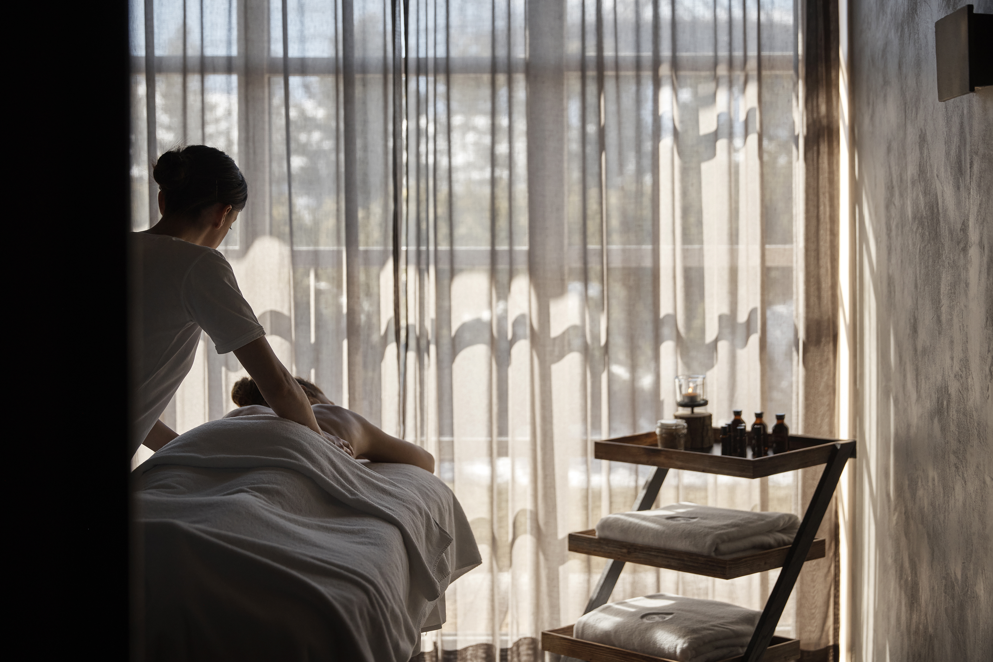 A luxurious spa with sheer curtains partially drawn over large windows, and massage oils set out on a wooden tray