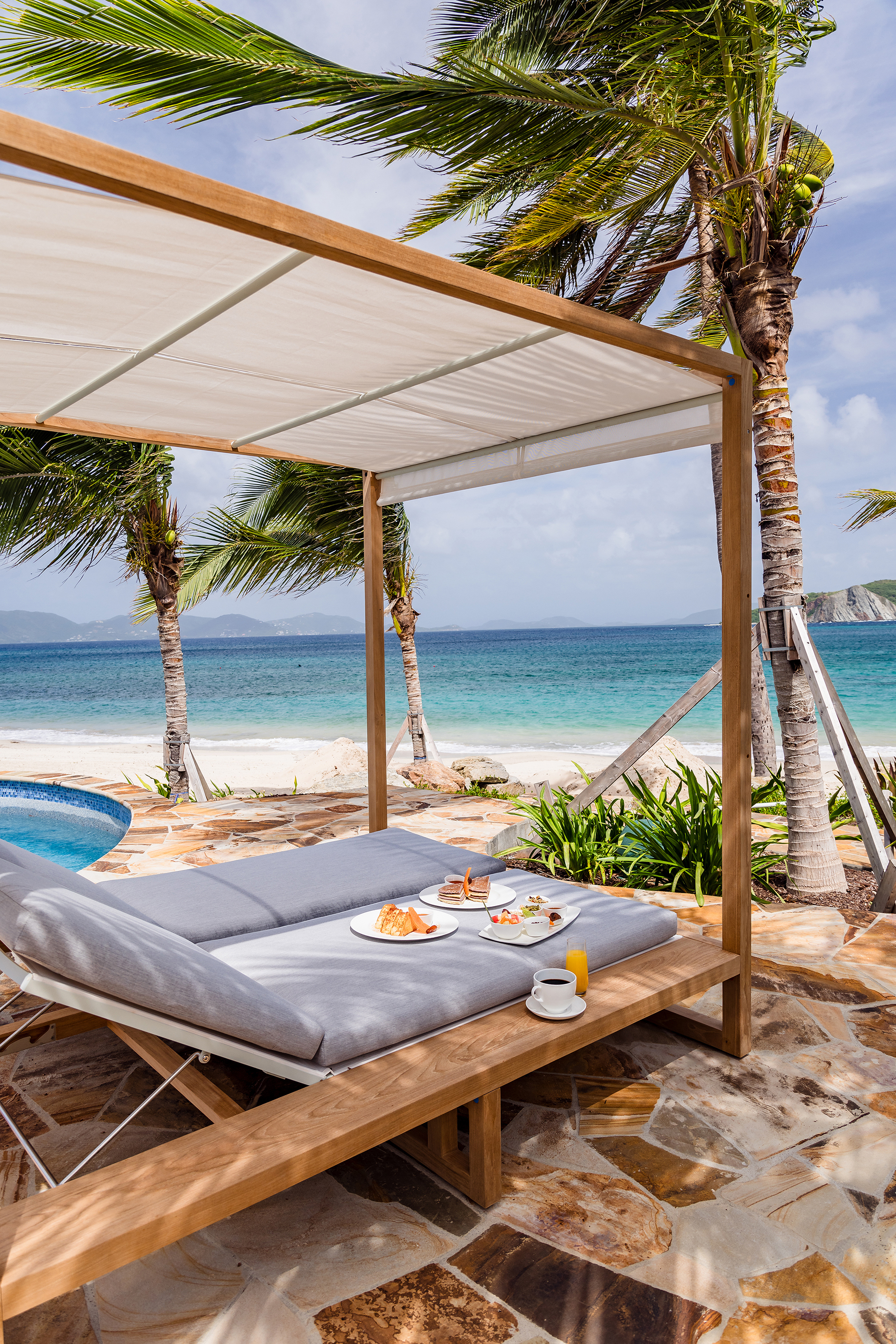 Wooden cabana above sun loungers laid with breakfast plates by a pool facing out to sea
