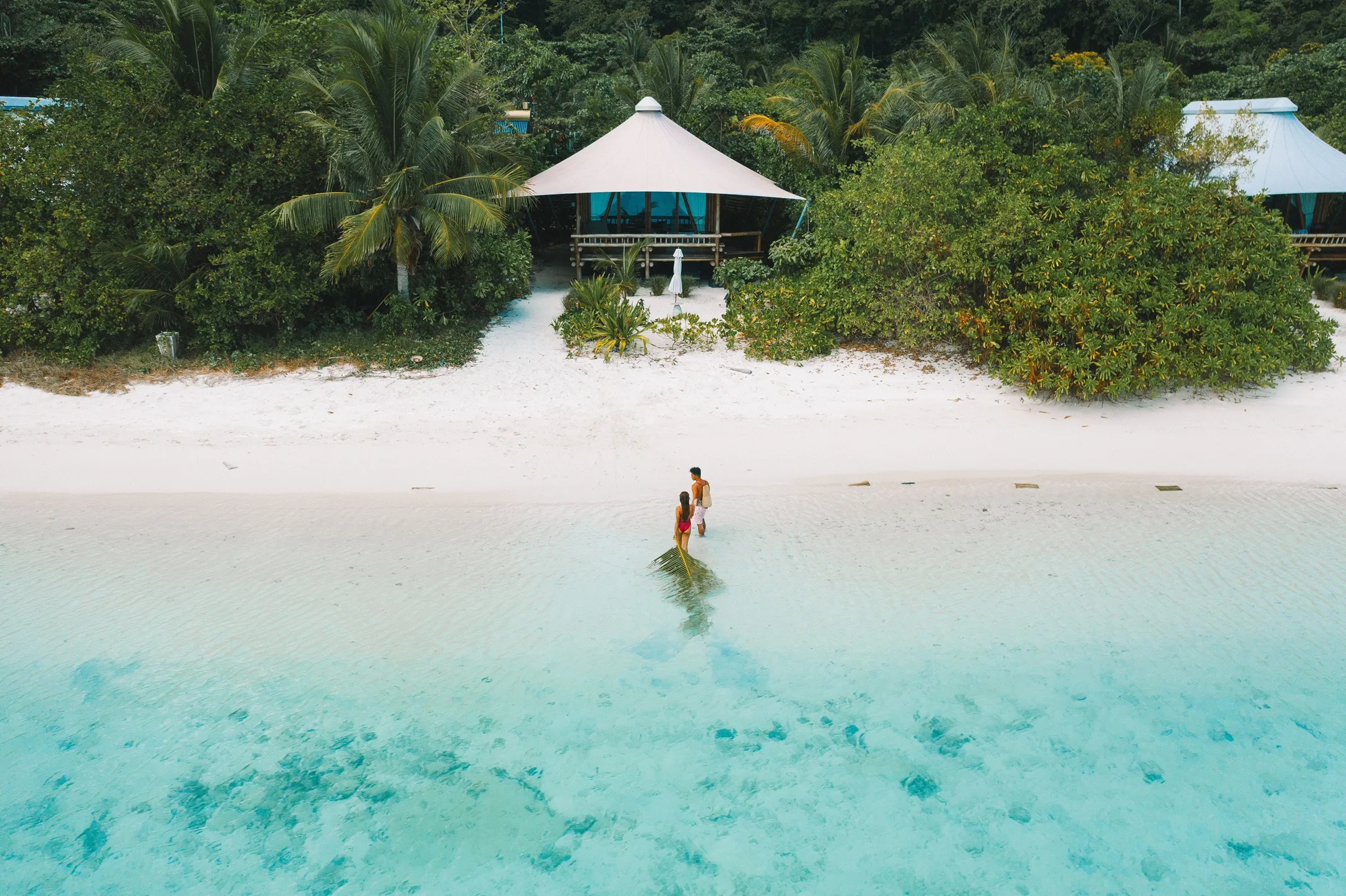 Couple standing in shallow turquoise water on a beach with greenery and a white tent in the background
