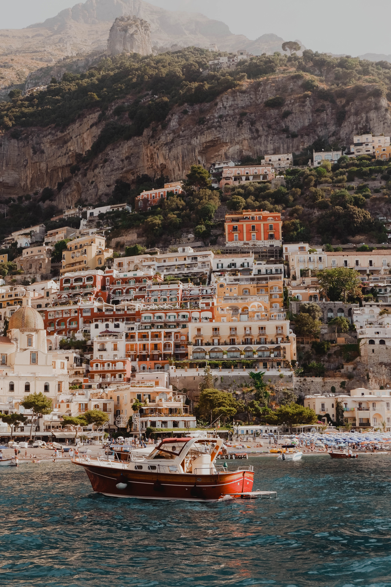 The cliffside village of Positano with different coloured buildings with the busy pebble beach and small boats in front