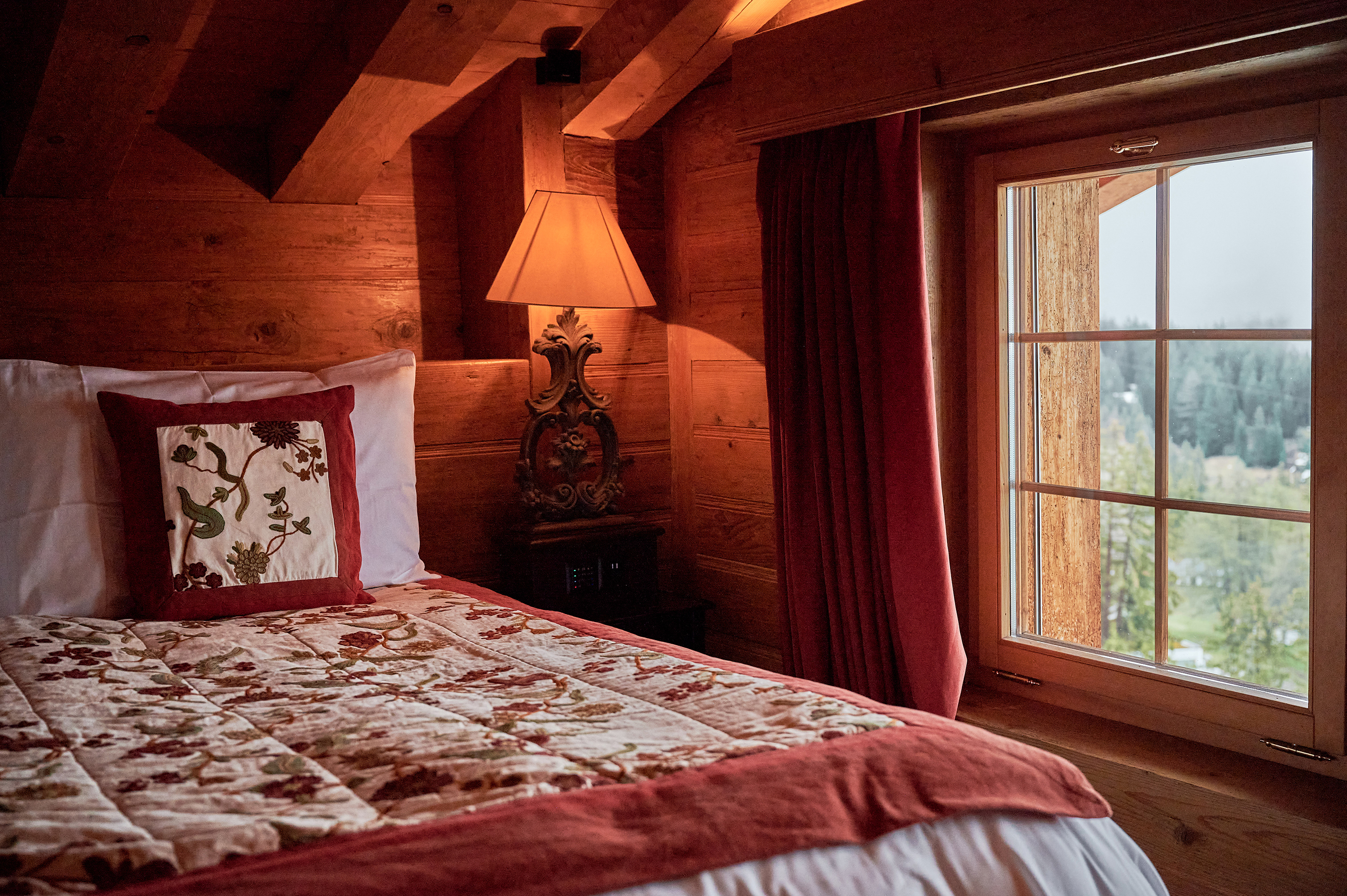 Details of a cosy suite at LeCrans featuring a bed with patterned cushions and throw, ornate lamp and a small window with a view of the landscape