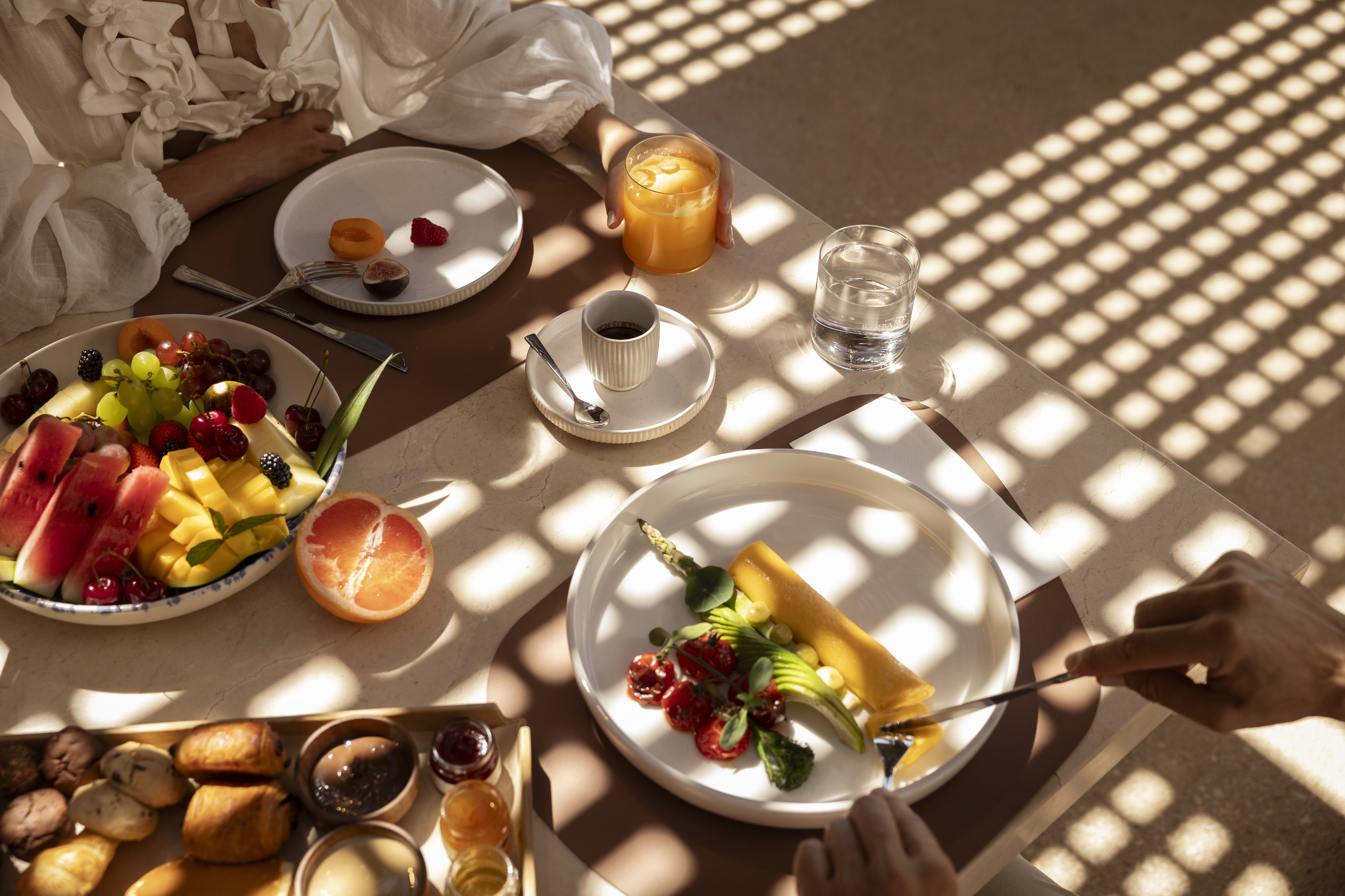 Fruit and breakfast pastries set on a table with two people ready to start dining under dappled sunlight