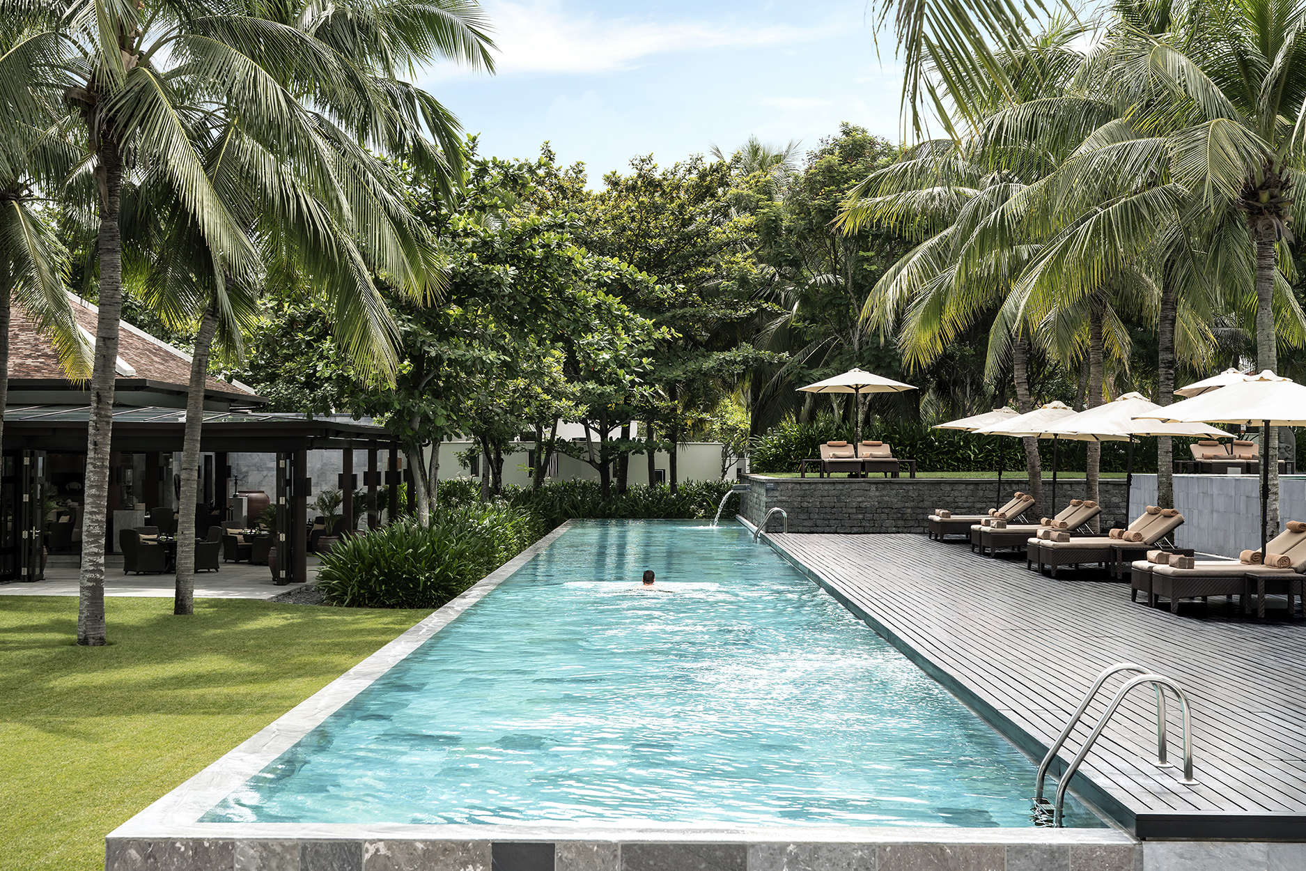 An infinity pool flanked by a terrace with sun loungers on one side and a lawn and trees on the other at Four Seasons Resort Nam Hai