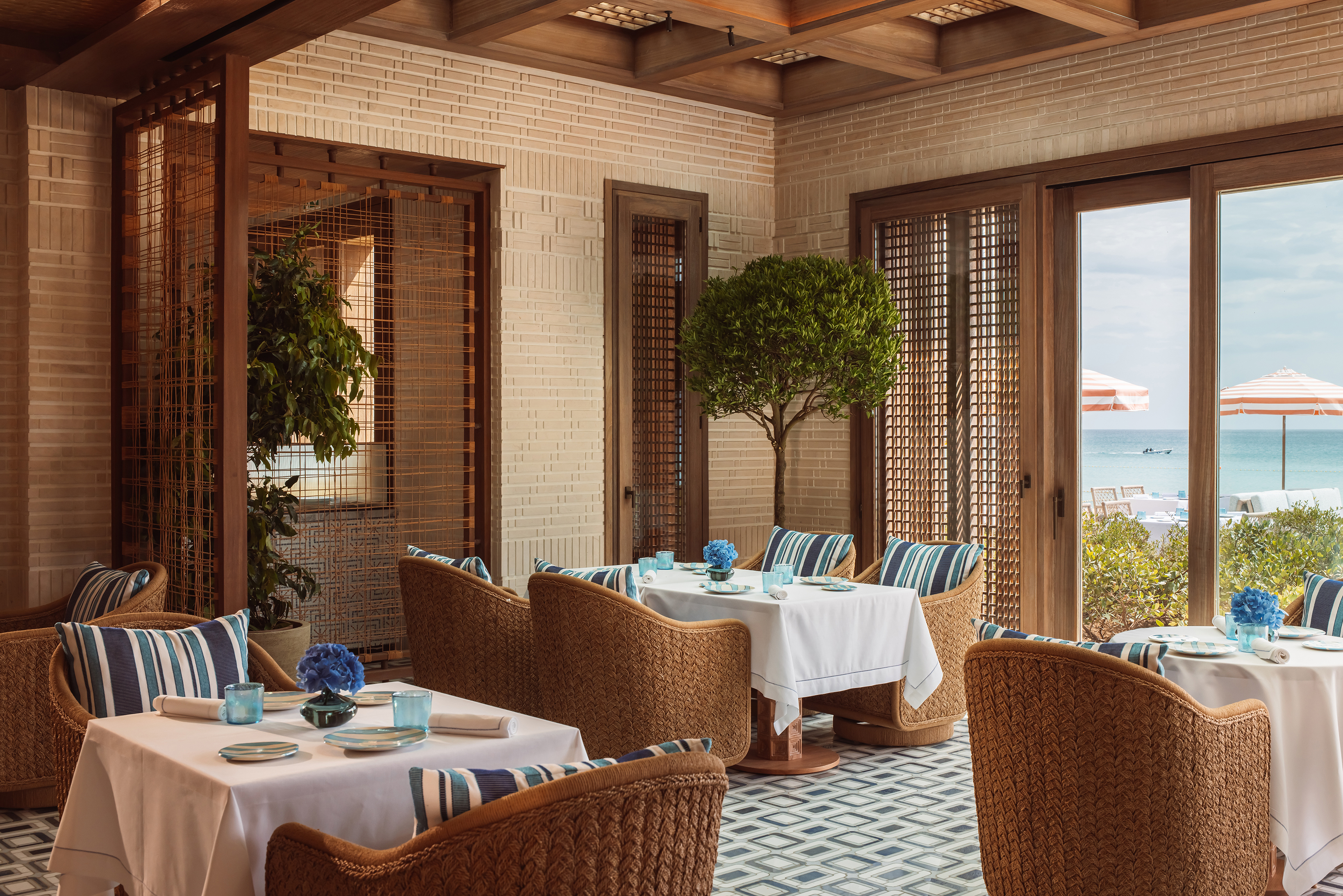 A comfy and airy restaurant featuring rattan furniture comfy striped cushions and lattice sun screens, with views of the ocean through the windows 