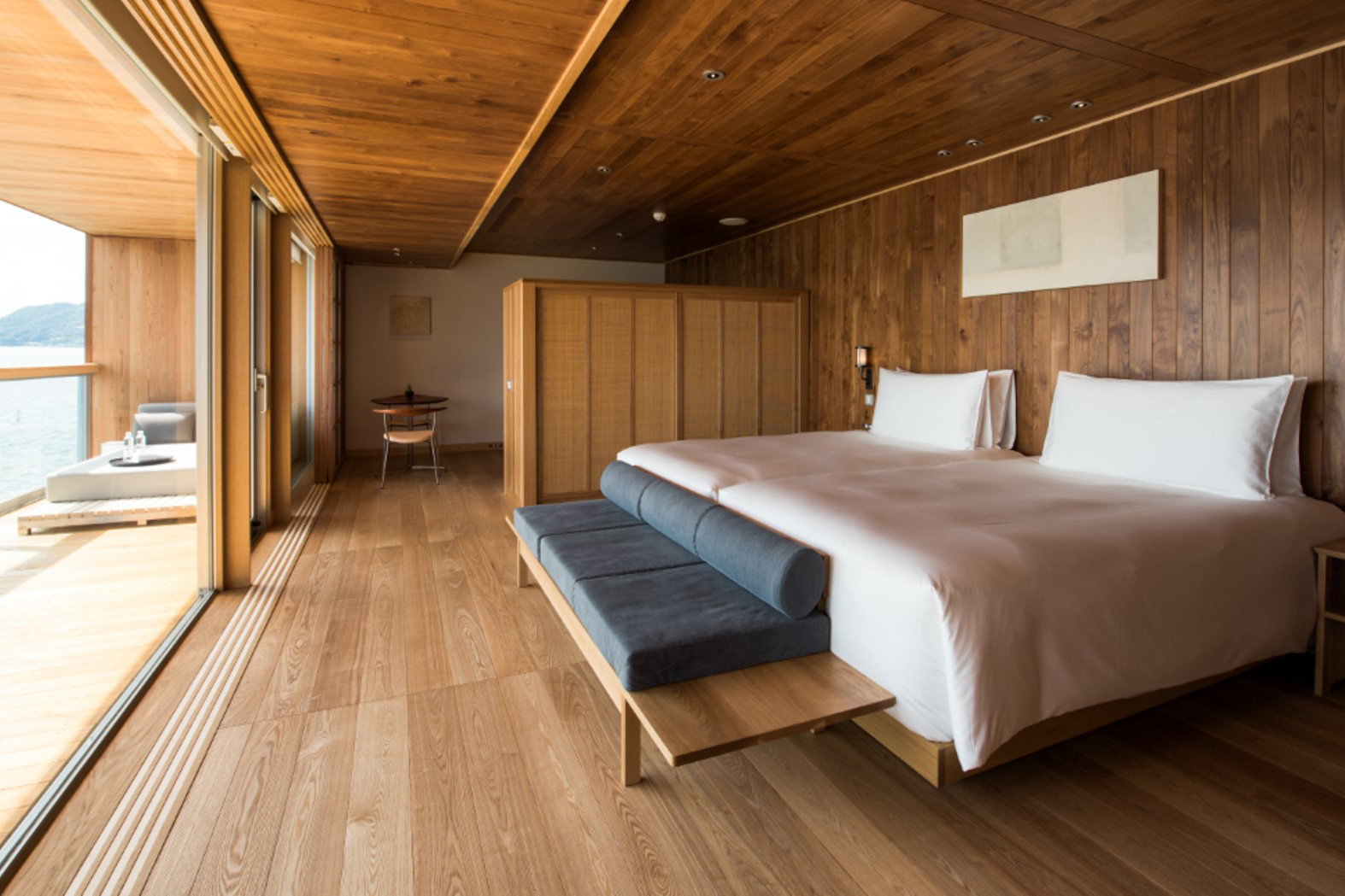 A wood-panelled bedroom featuring a large bed, ottoman and wooden screen, with a glimpse of a terrace through the large windows