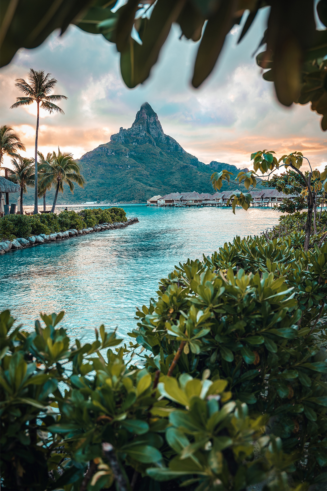 A scenic view of a mountain peak seen through foliage, with overwater bungalows and a tranquil sea at sunset.