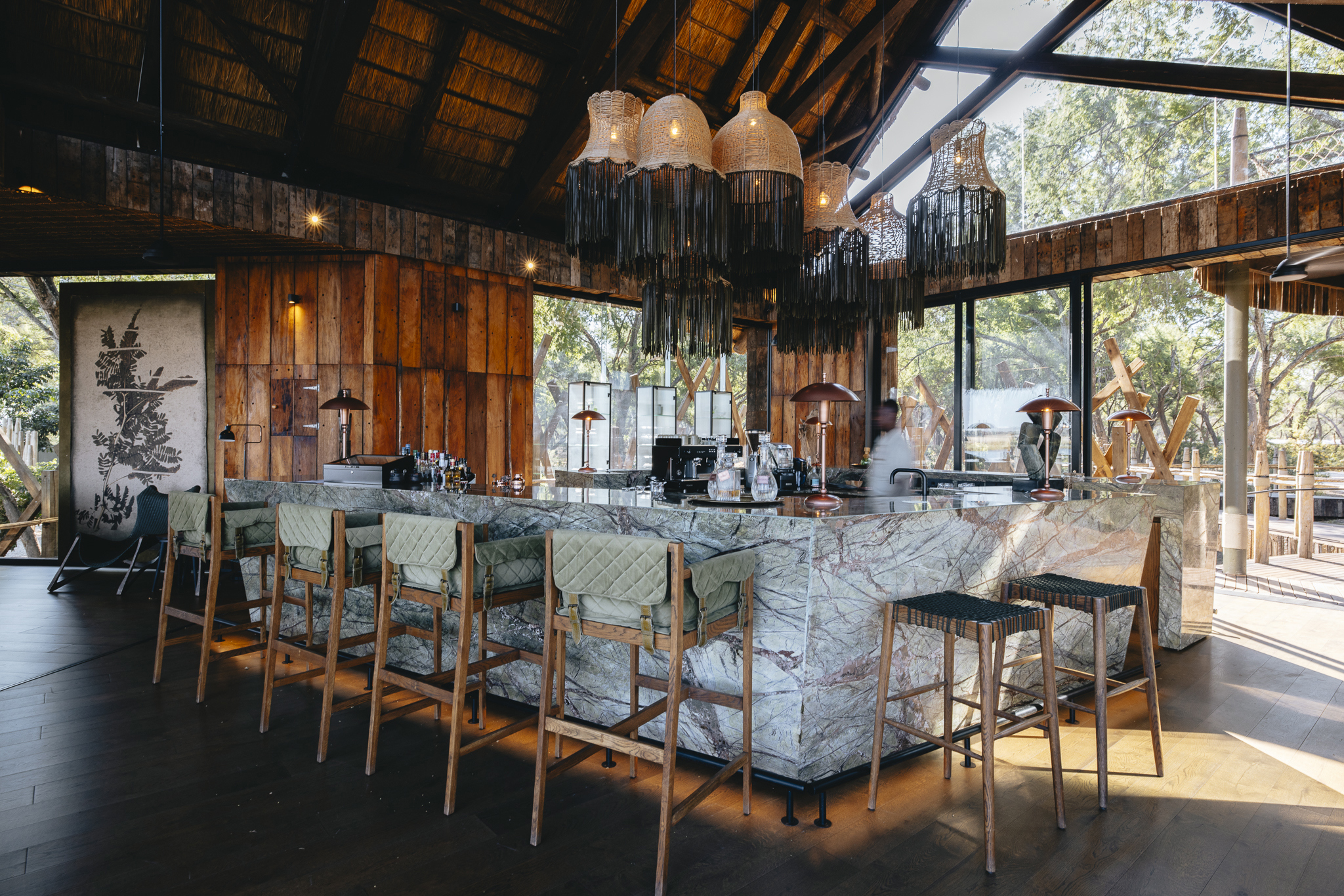 A large square marble bar with stools beneath ornate light shades in a thatch and glass building 