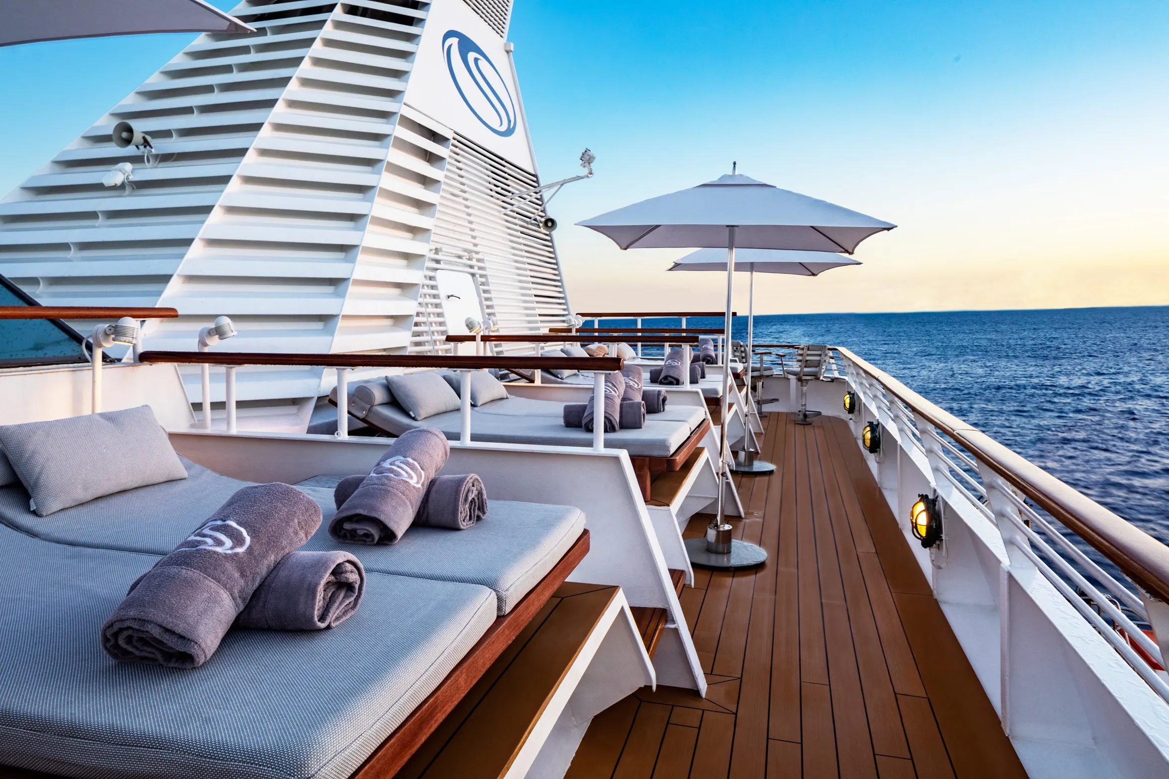Cruise ship deck with cushioned lounge chairs, white umbrellas, and ocean views at sunset.