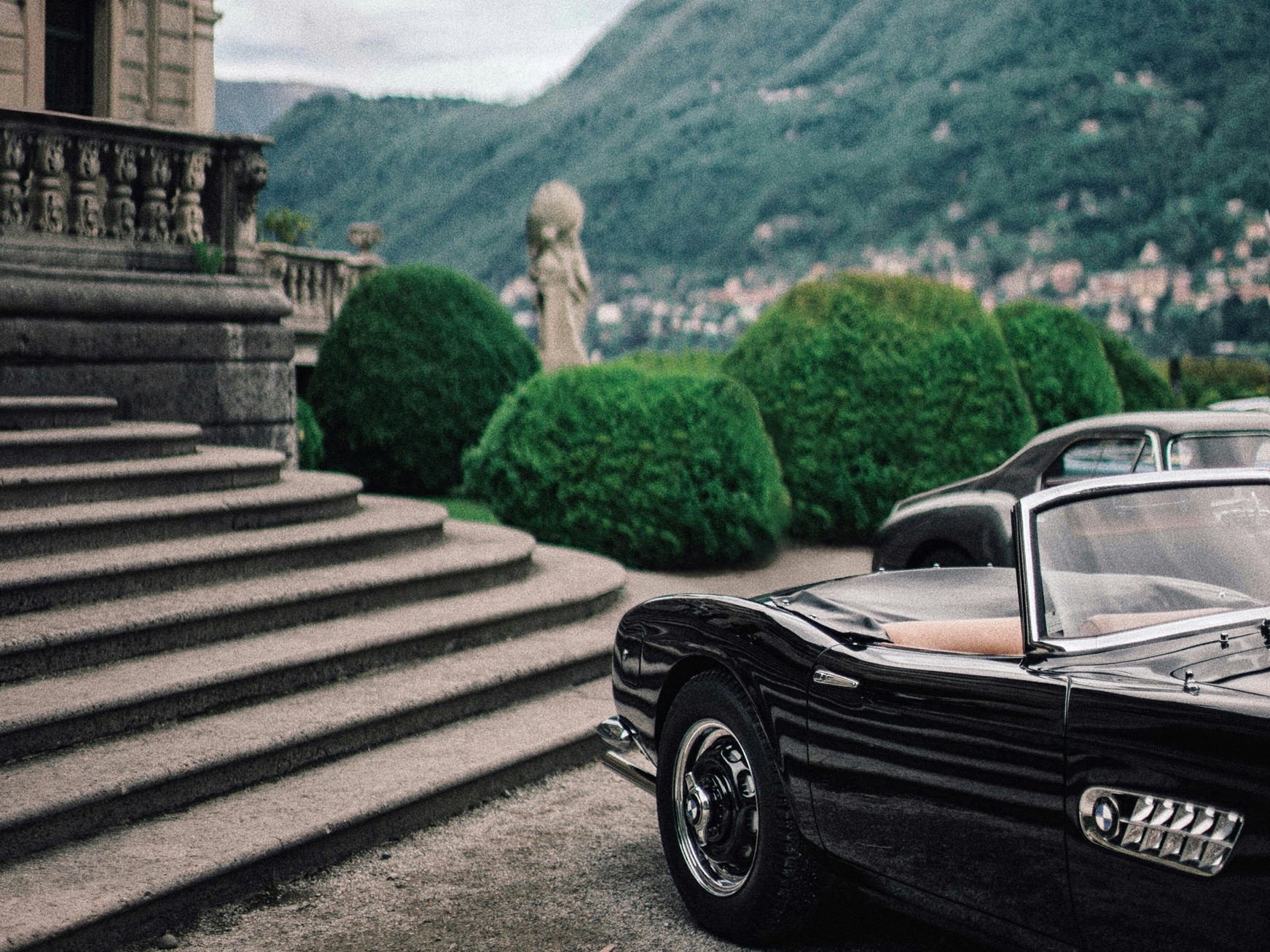 Black convertible parked in front of a grand stone staircase with manicured bushes and mountains in the background