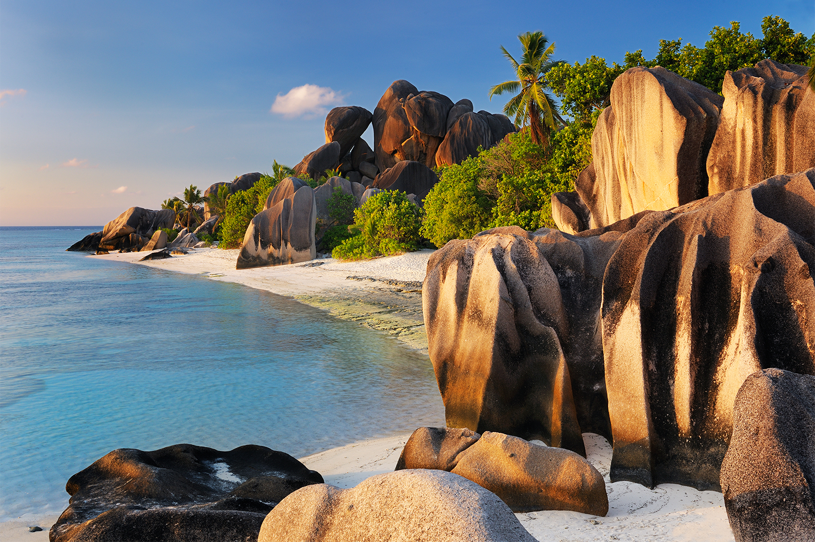 A tropical beach with distinctive large granite boulders, clear blue water, and lush green palm trees under a partly cloudy sky.