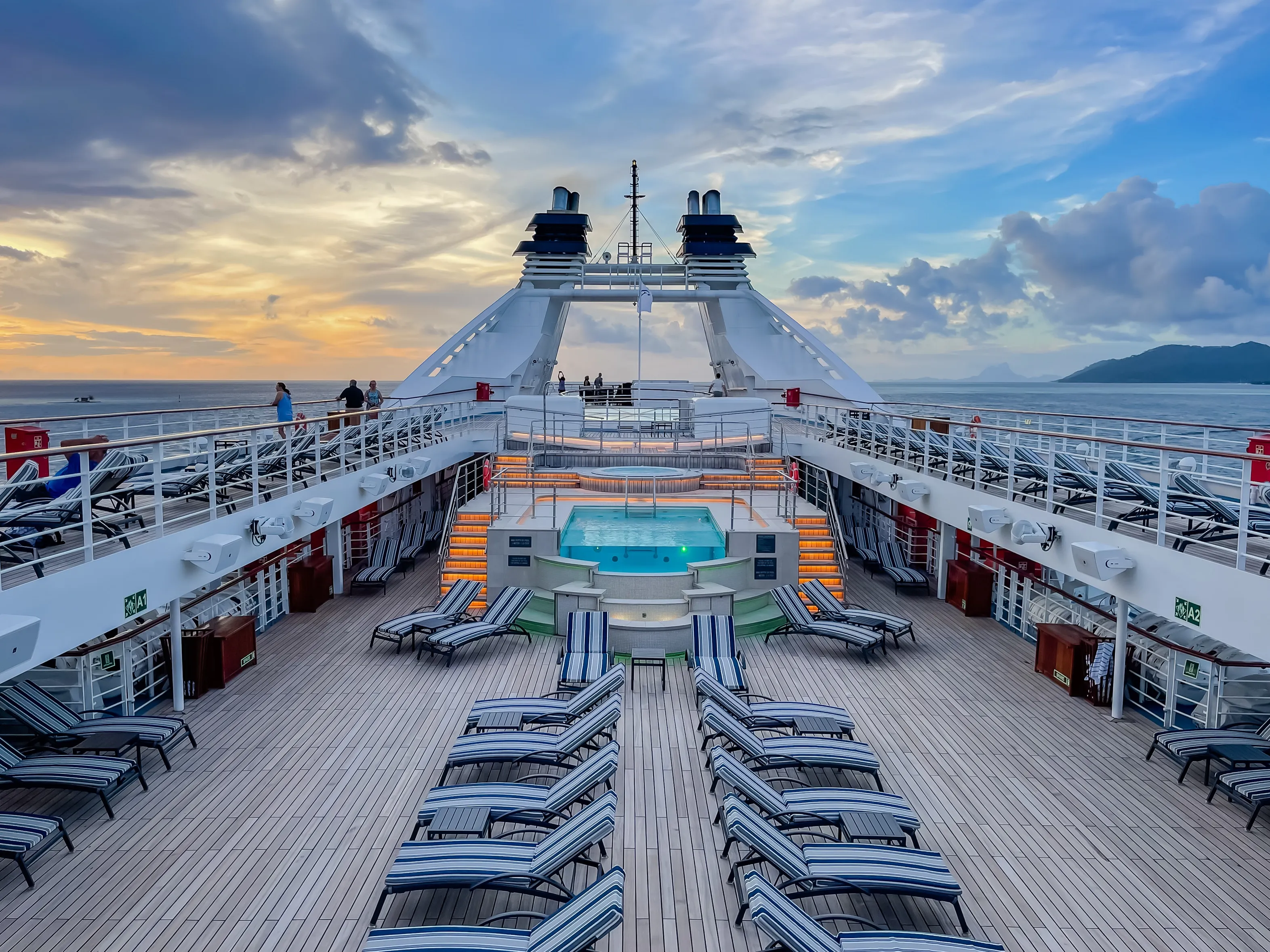 A cruise ship deck with lounge chairs around a central pool, under a partly cloudy sunset sky.