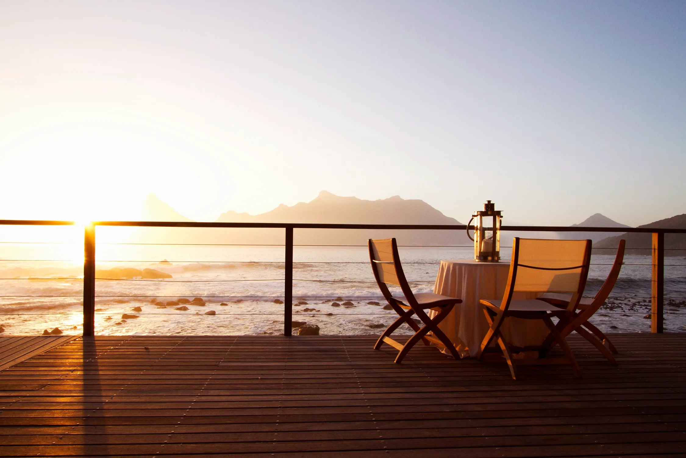 Small table with two chairs set on a wooden deck overlooking the ocean at sunset, with mountain silhouettes in the background.