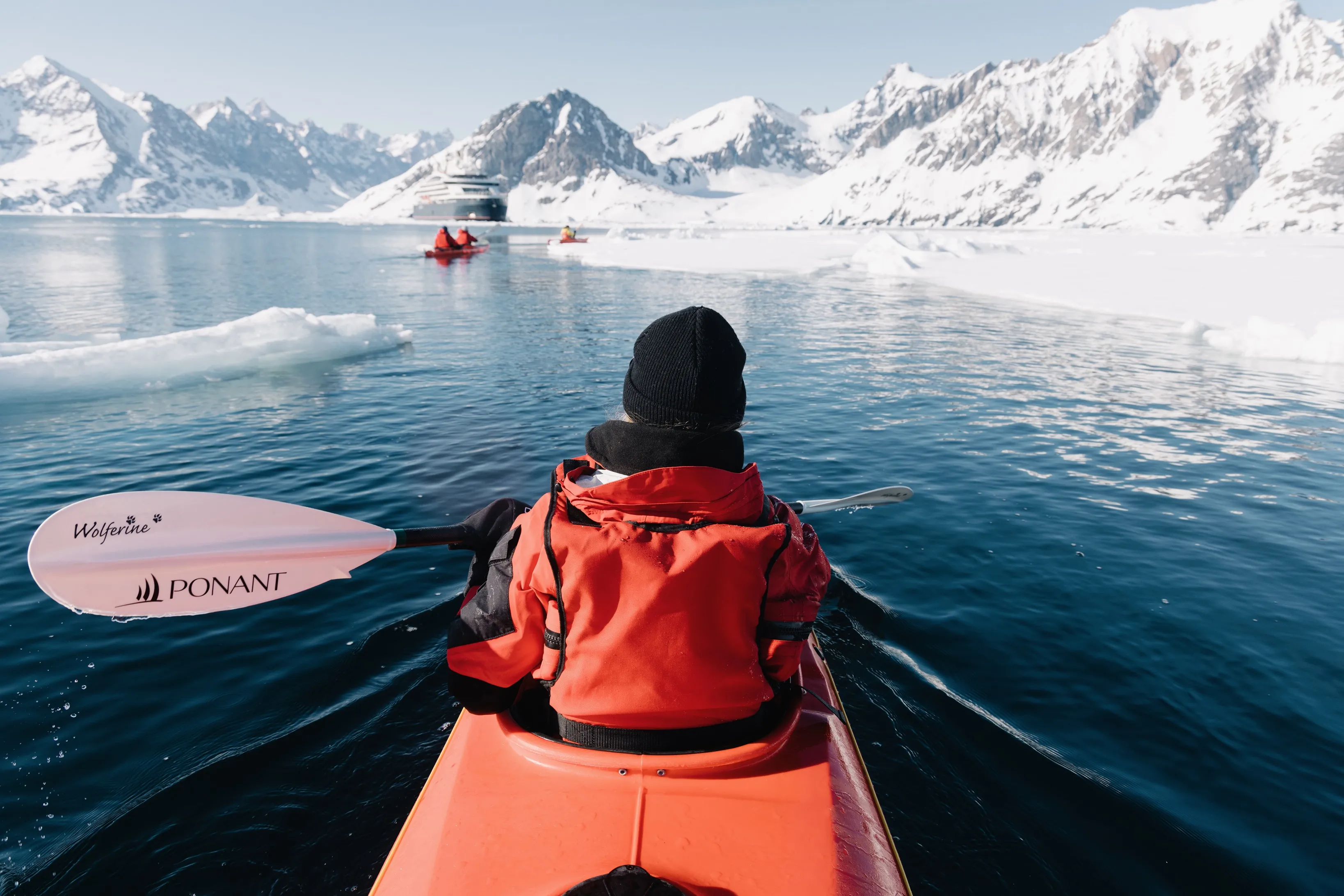 Ponant guest kayaking through icy waters with snow-covered mountains and fellow kayakers in the distance.