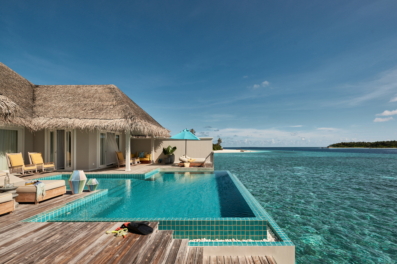 The side of a pool deck in a Water Villa at Finolhu with a large infinity pool backed by sun loungers and a thatched building with a view of the ocean beyond