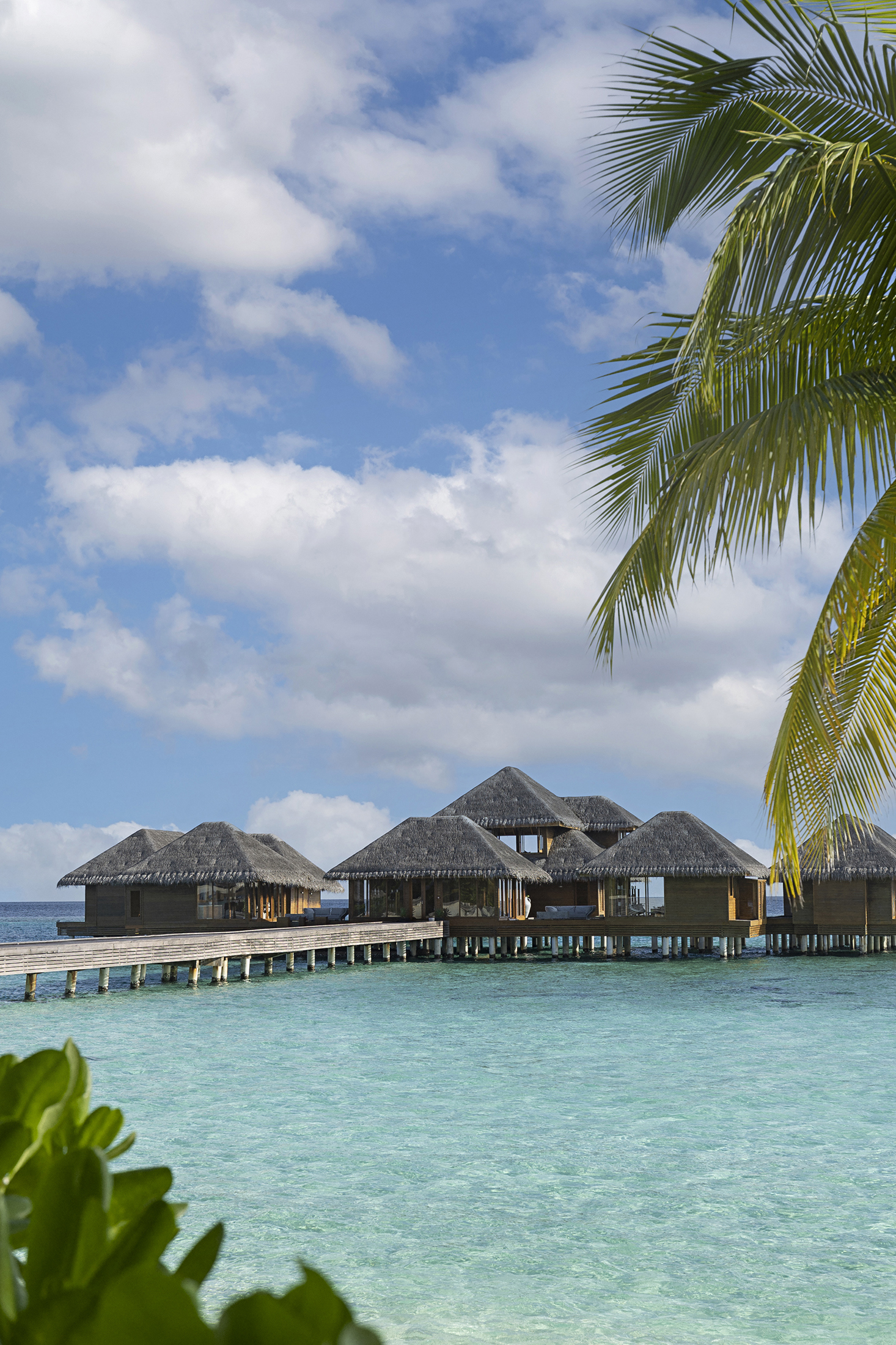A view from the beach of the overwater spa at Huvafen Fushi through greenery featuring a wooden walkway leading to a cluster of thatched buildings