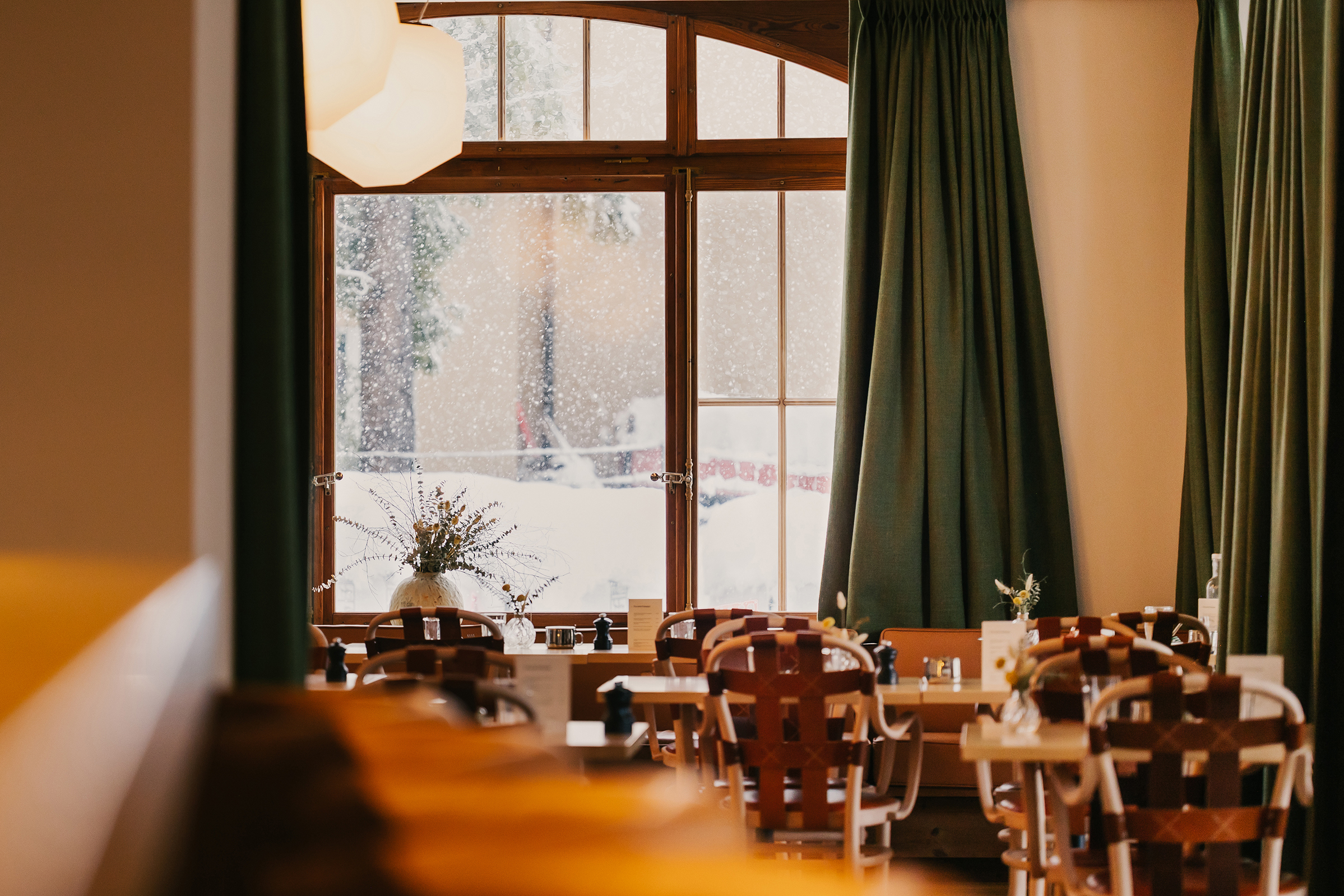 Cosy tables and chairs set near a large window with green curtains and a snowy scene outside