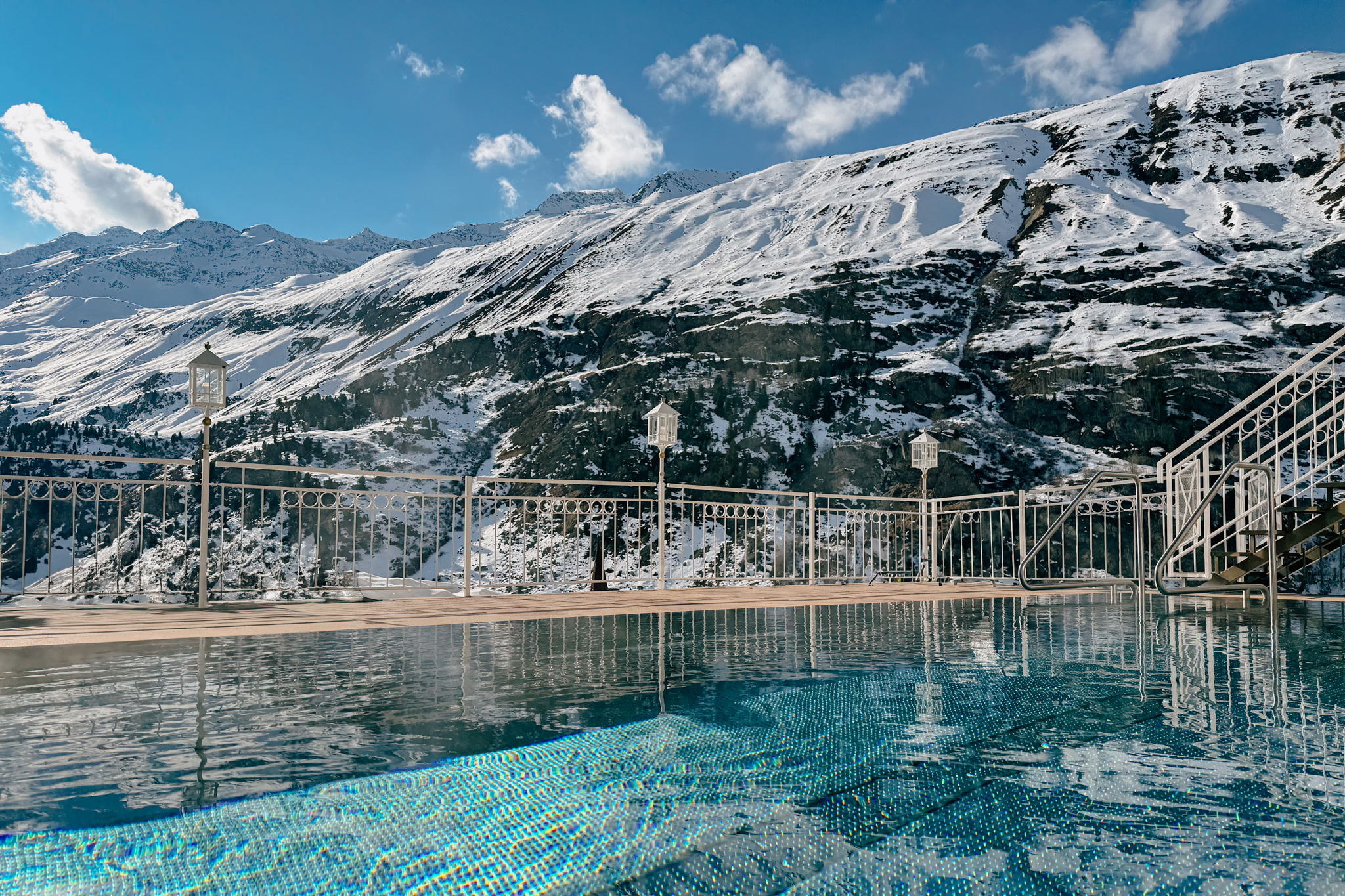 An outdoor pool with clear blue water overlooking snowy mountains under a bright blue sky at the Gourmet and Wine Hotel Austria