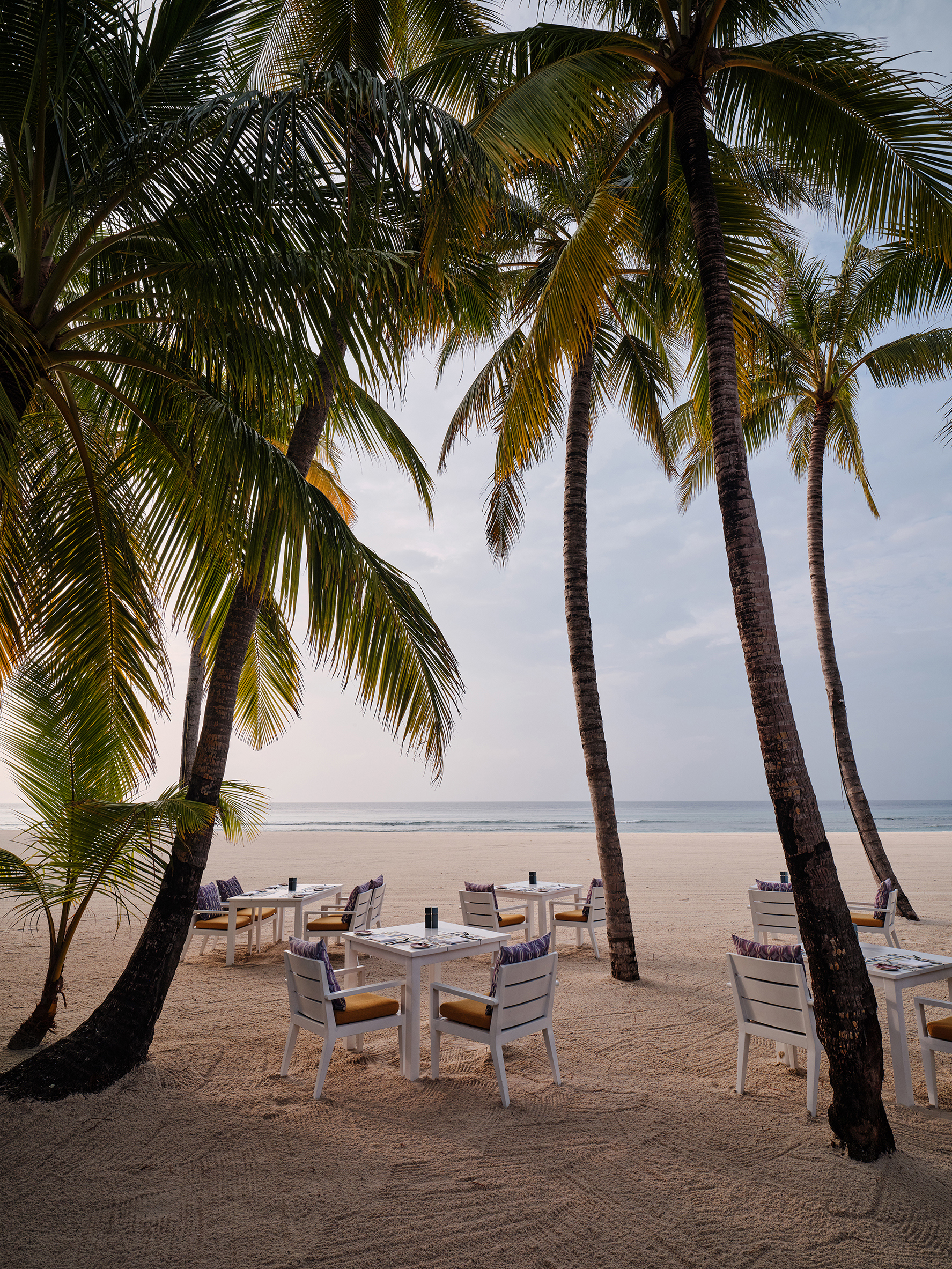 Five white tables and chairs set for a meal on the sand between palms