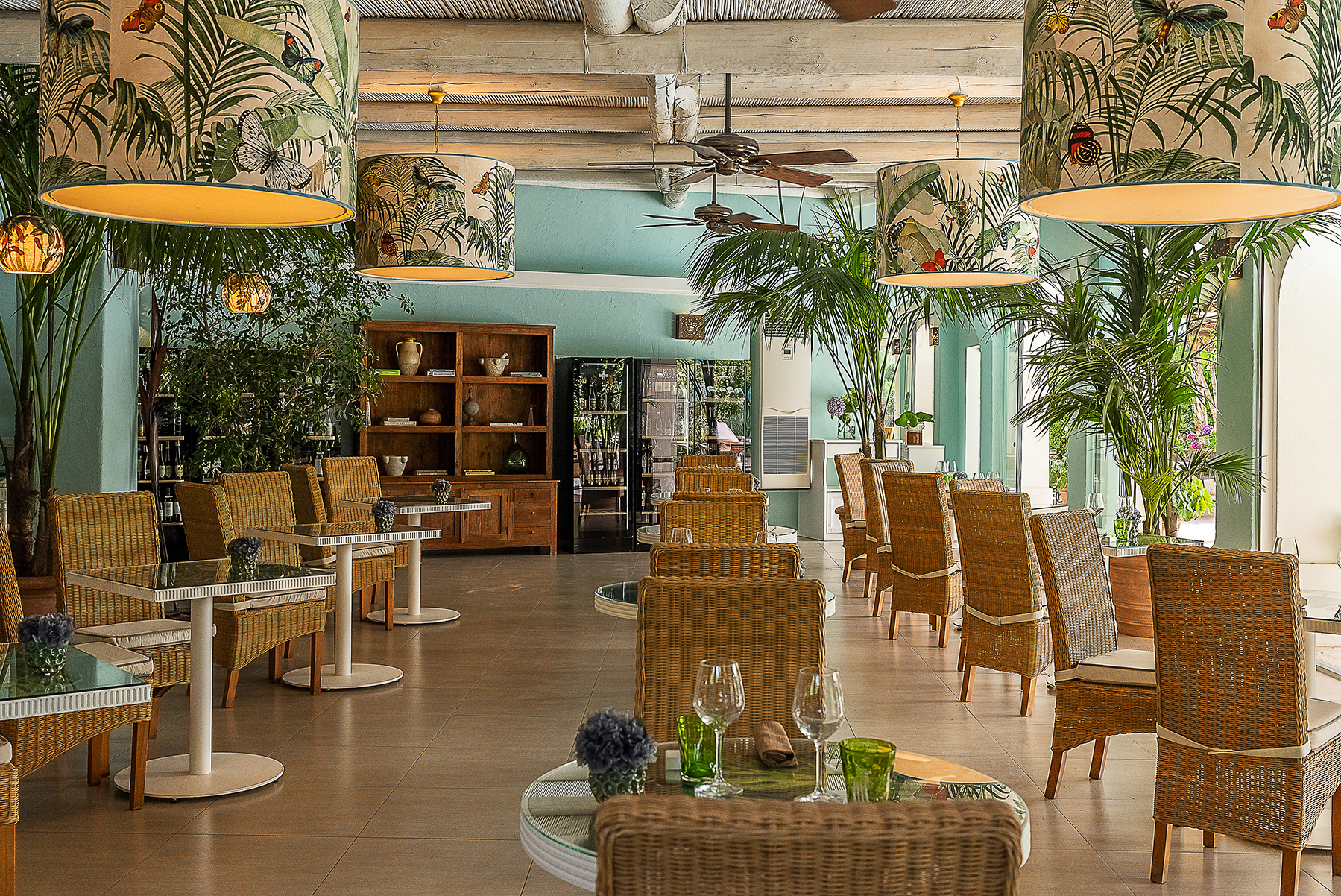 Elegant dining area in Il Corbezzolo Restaurant at Botania Relais & Spa in Ischia featuring wicker chairs, tropical-themed lampshades, and lush greenery.