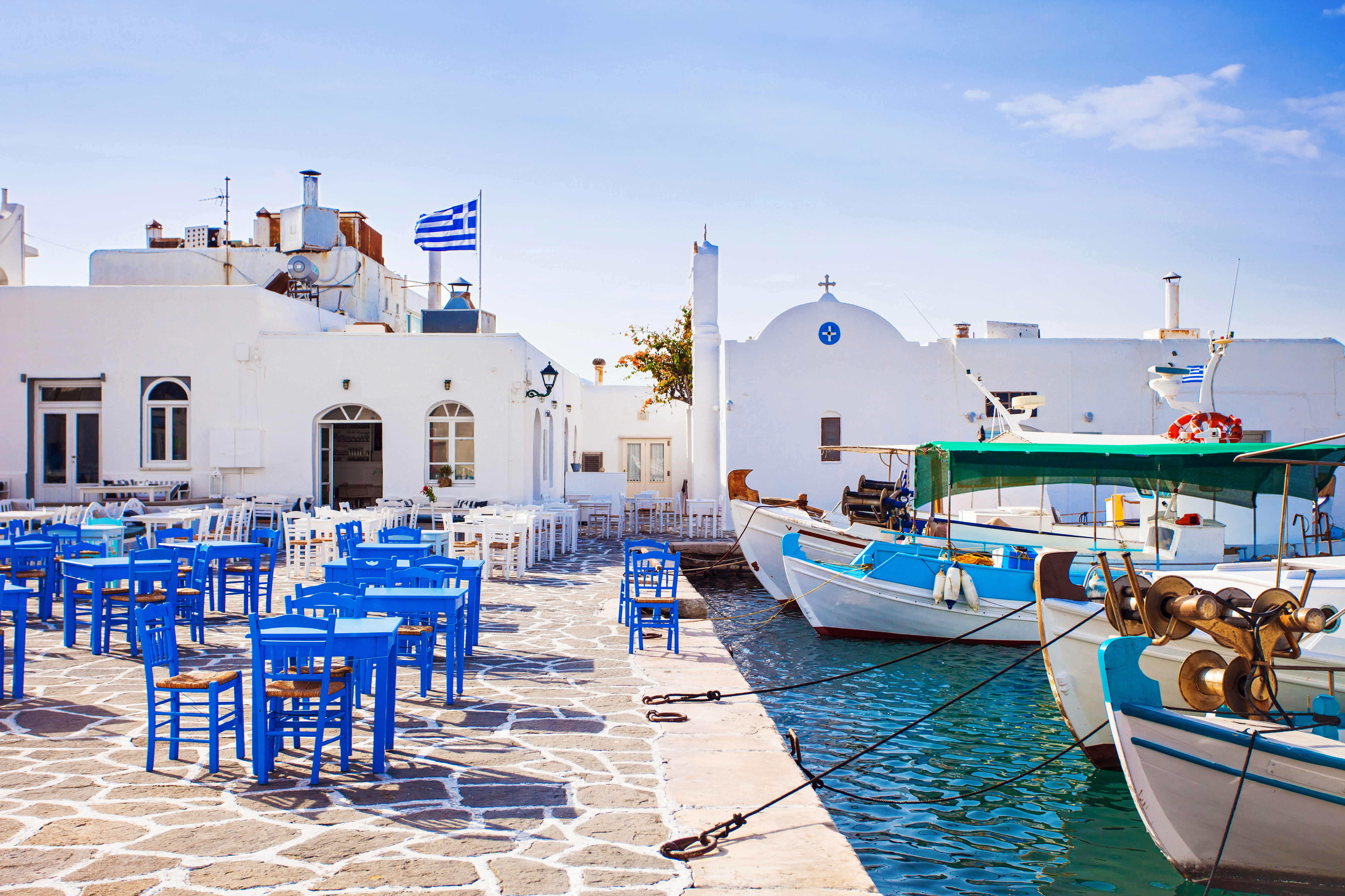 Cobbled marina on Paros set with blue dining tables and boats moored up in front of whitewashed buildings
